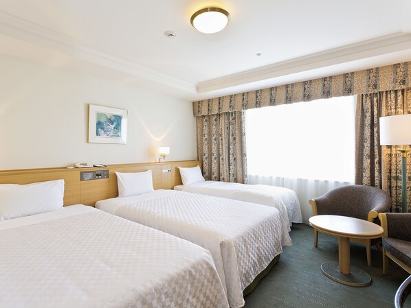 A hotel room with two beds, a small table, a chair, a lamp, a painting, and a large window with curtains.