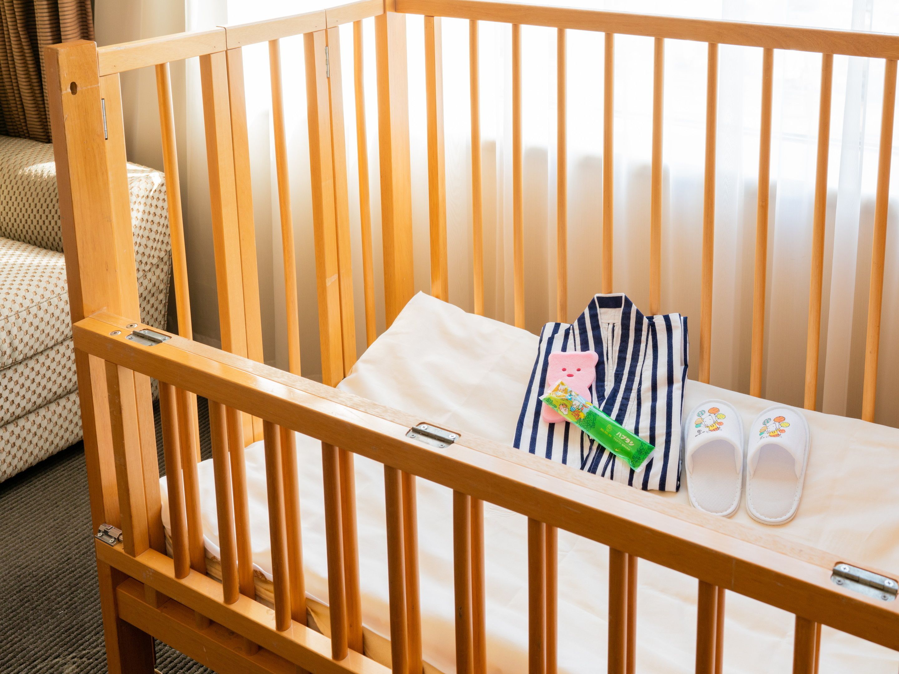 A wooden crib with a mattress, a striped pillow, a green bottle, and a pair of slippers.
