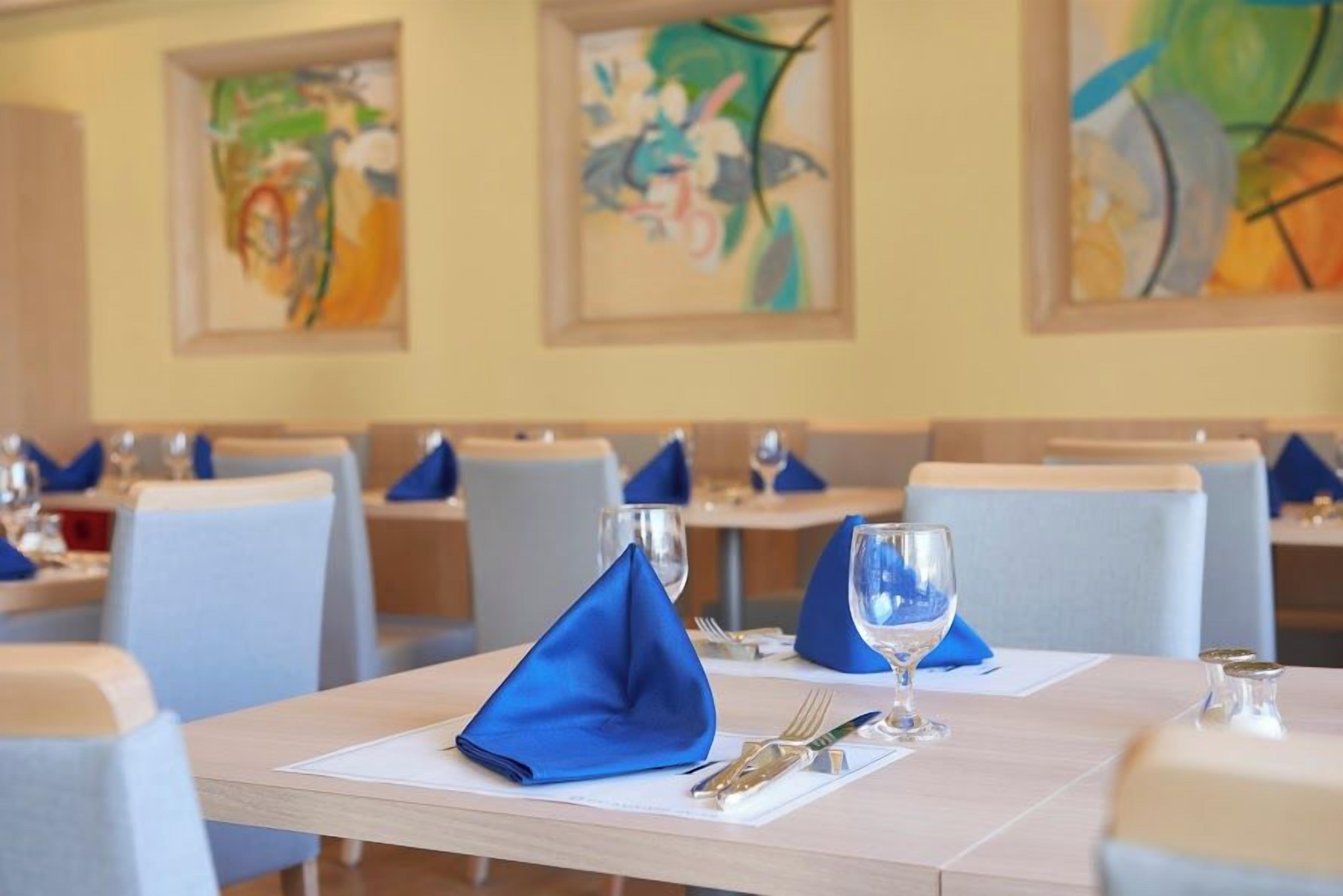 A dining area with tables set for two, each with a folded blue napkin, a glass, and cutlery.