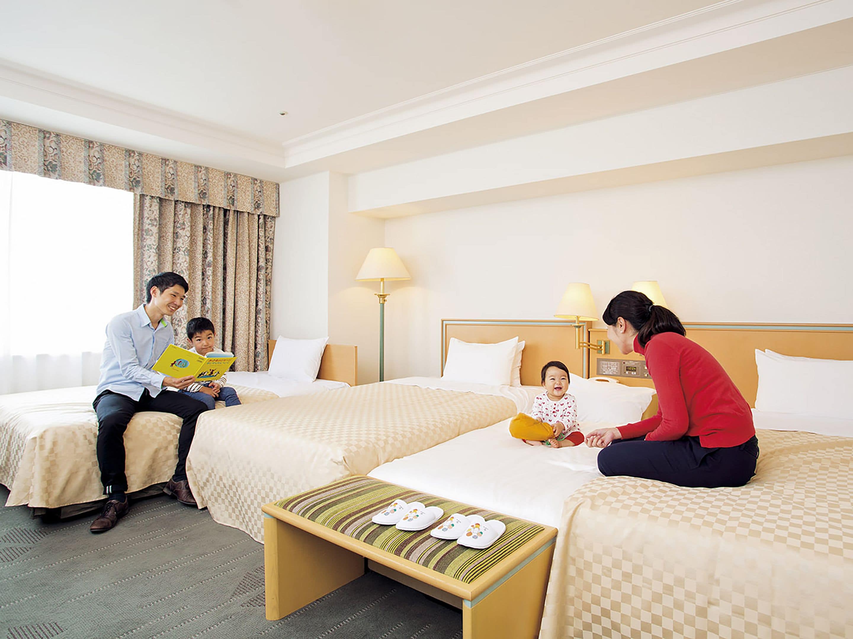 A hotel room with two beds, a bedside table with slippers, and a lamp.