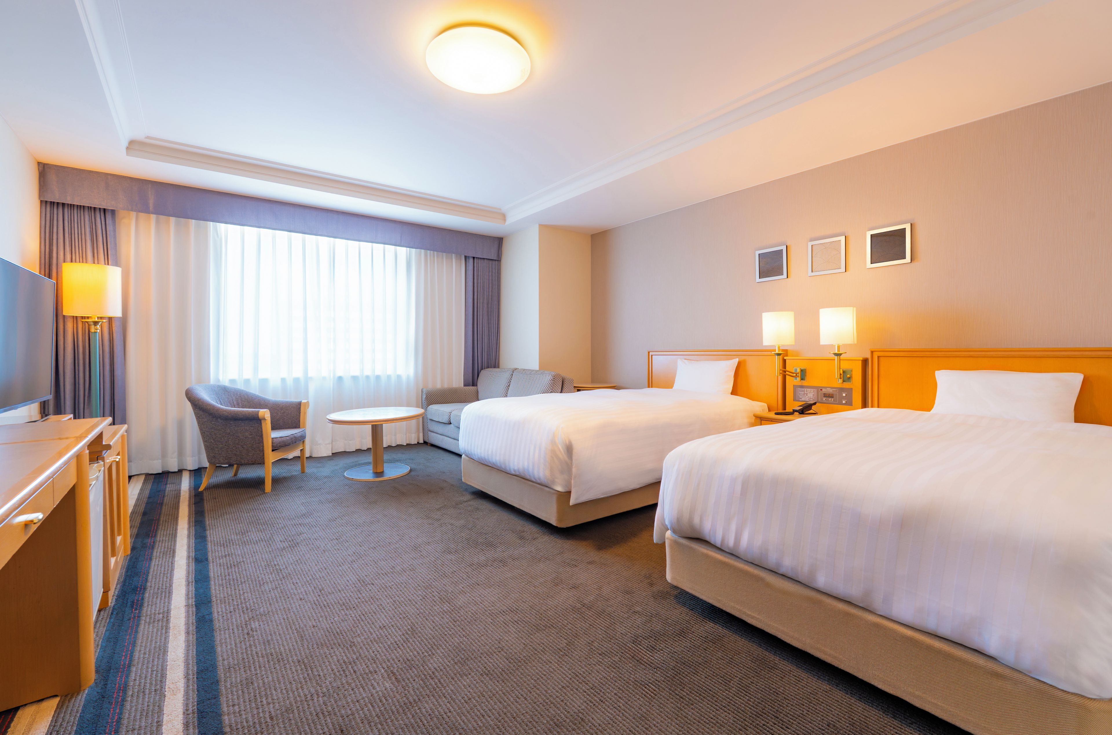 A hotel room with two beds, a desk, a chair, a small table, and a television.