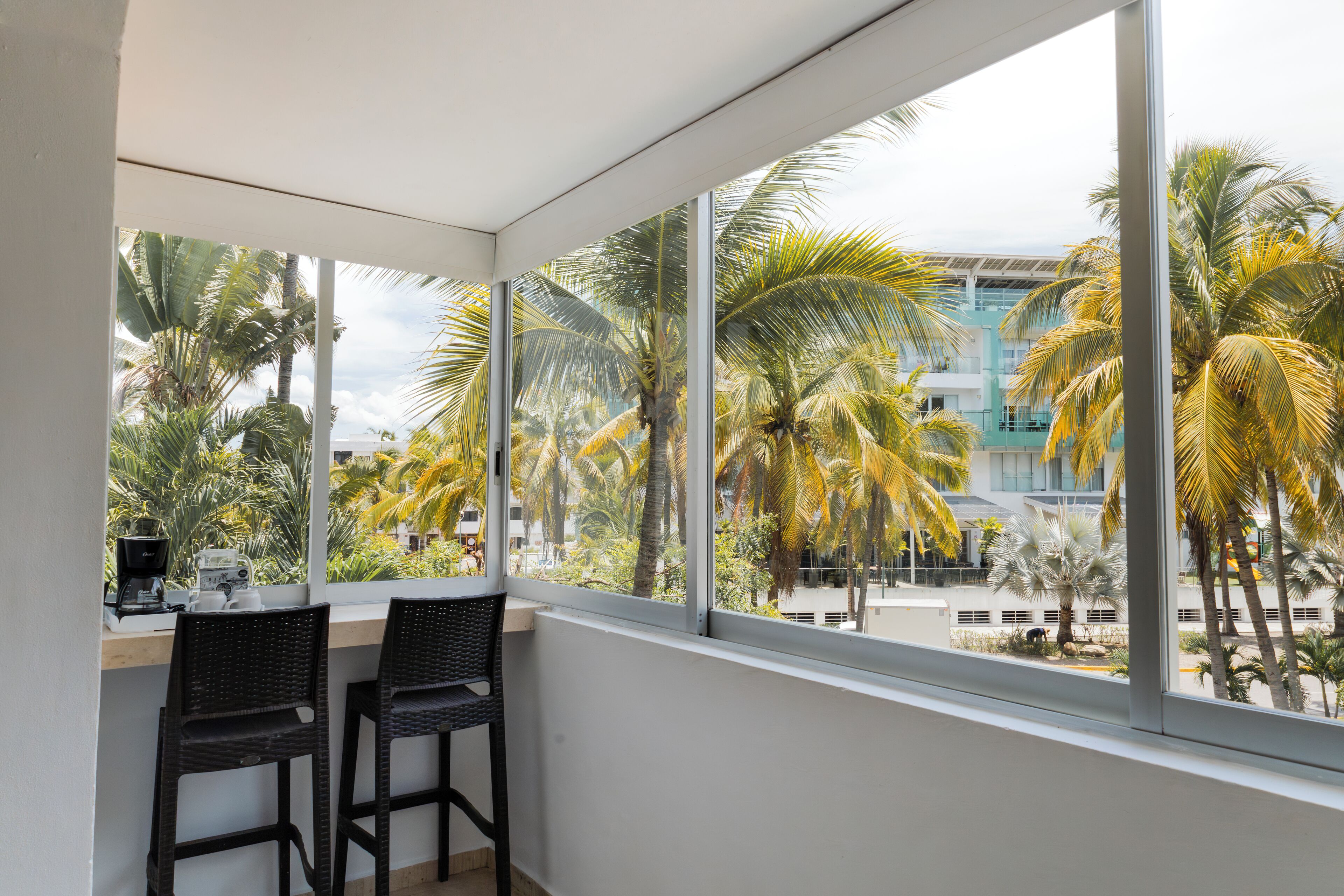 A balcony with two wicker chairs, a coffee maker, and a view of palm trees and buildings.