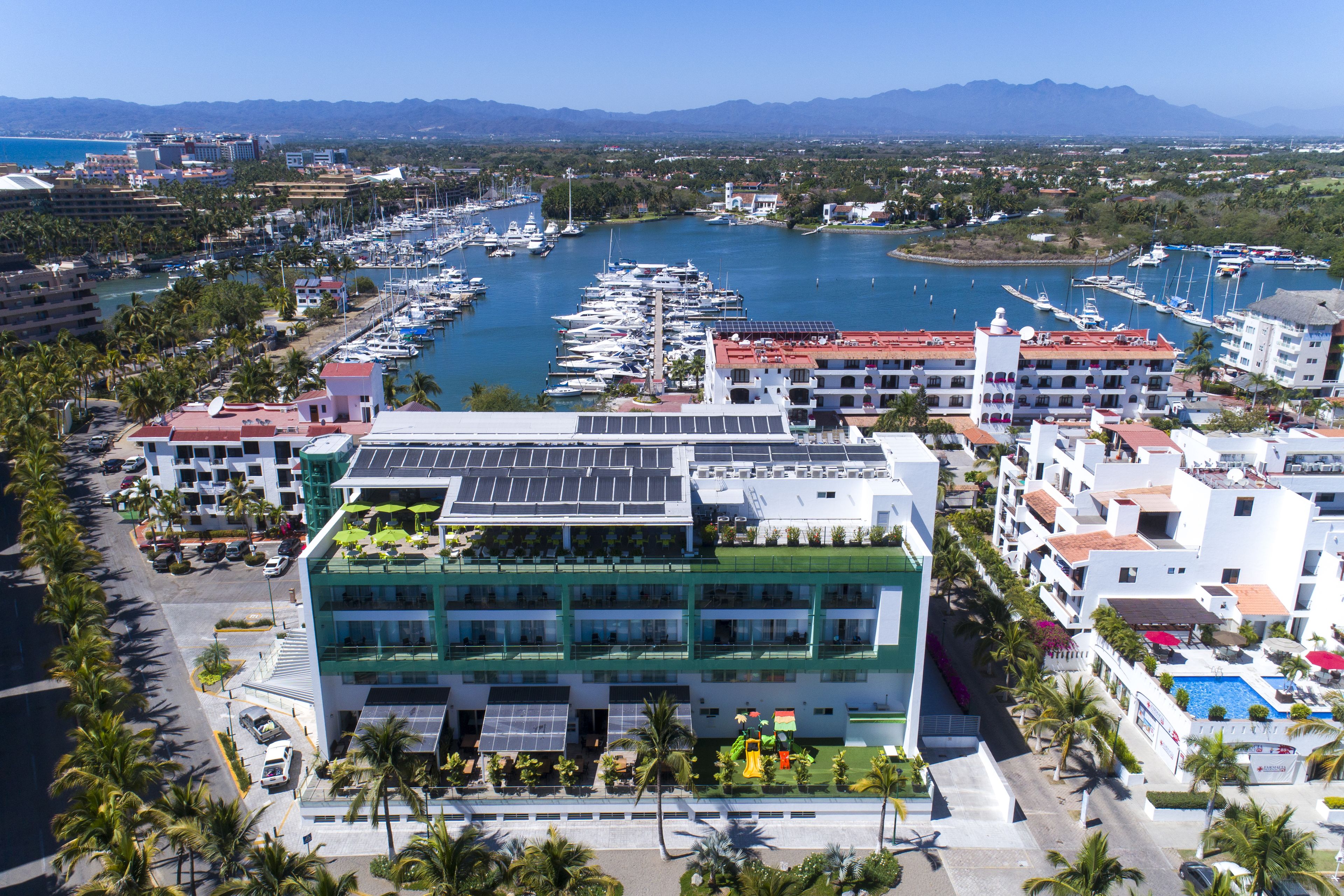 Un hotel frente al mar con varios pisos, un puerto deportivo con barcos y vistas al mar y a las montañas.