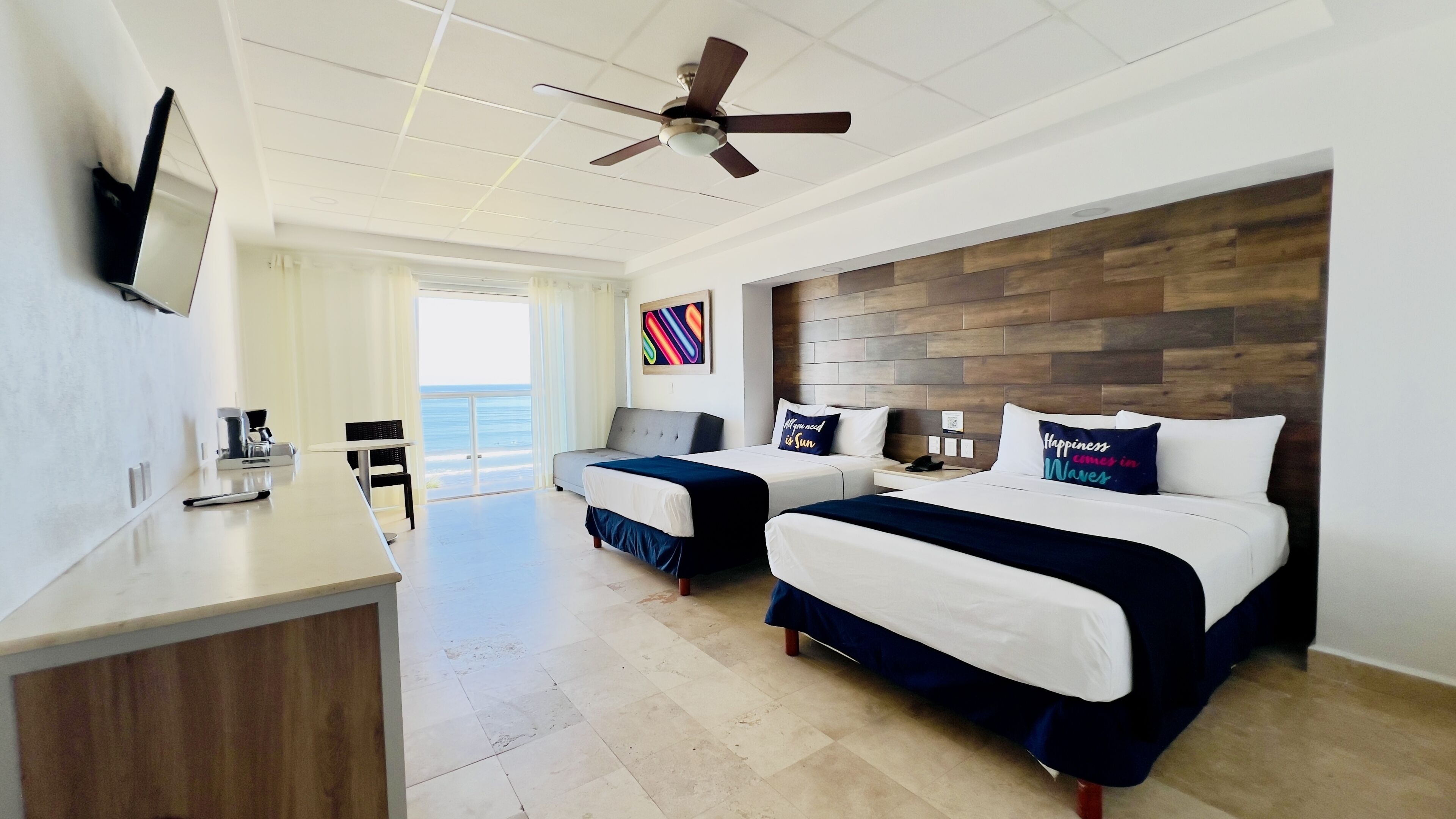 A hotel room with two beds, a desk, a chair, a ceiling fan, and a view of the ocean.