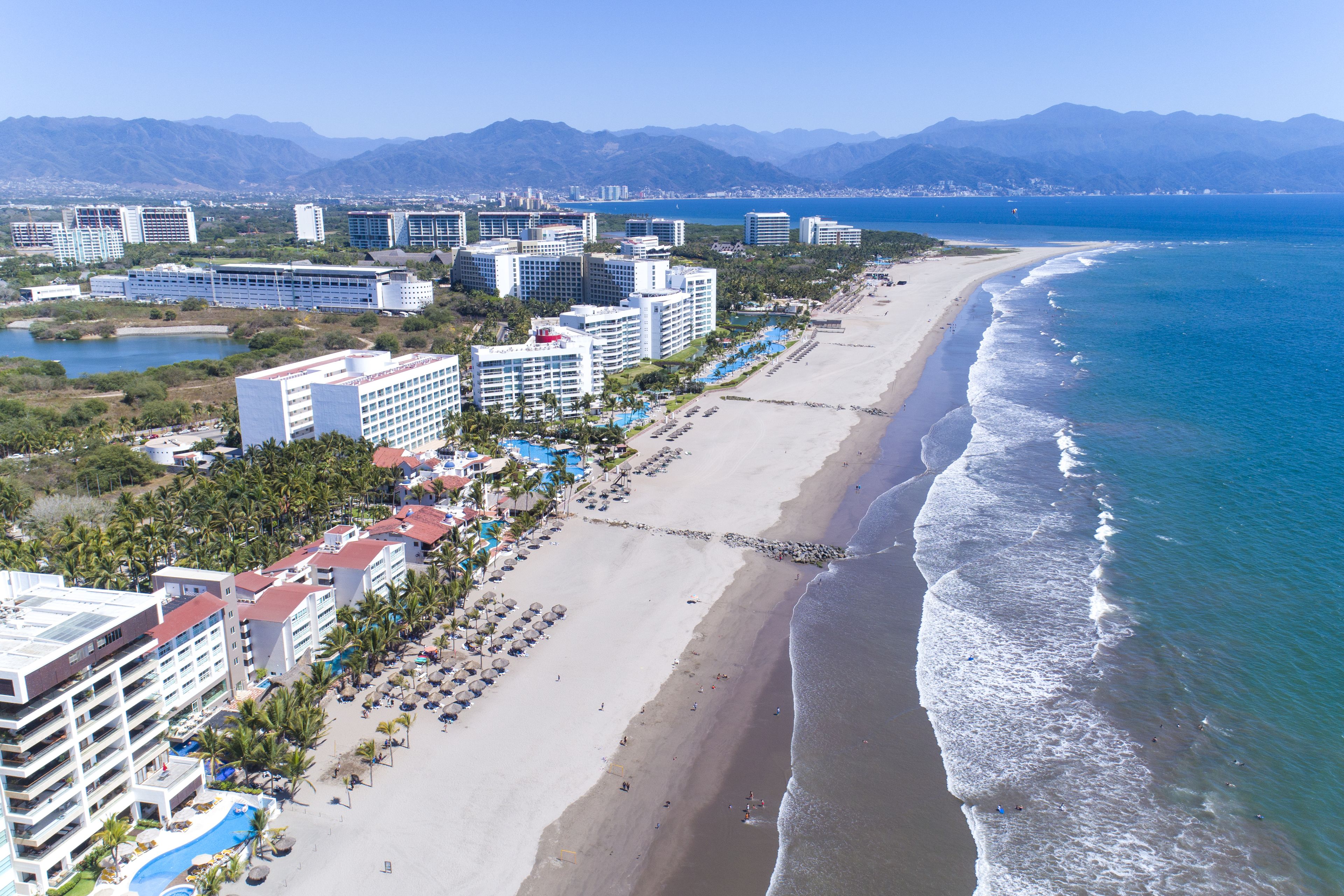 Ett hotell vid kusten med flera byggnader, en strand och en bergskedja i bakgrunden.