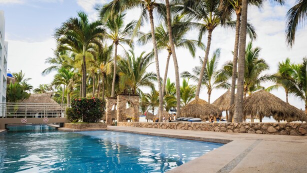 A swimming pool surrounded by palm trees and thatched-roof structures. - Villa Varadero (Nuevo Nayarit)