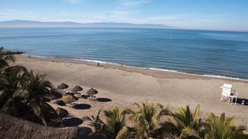 Una playa con palmeras, sombrillas de paja y algunas sillas.