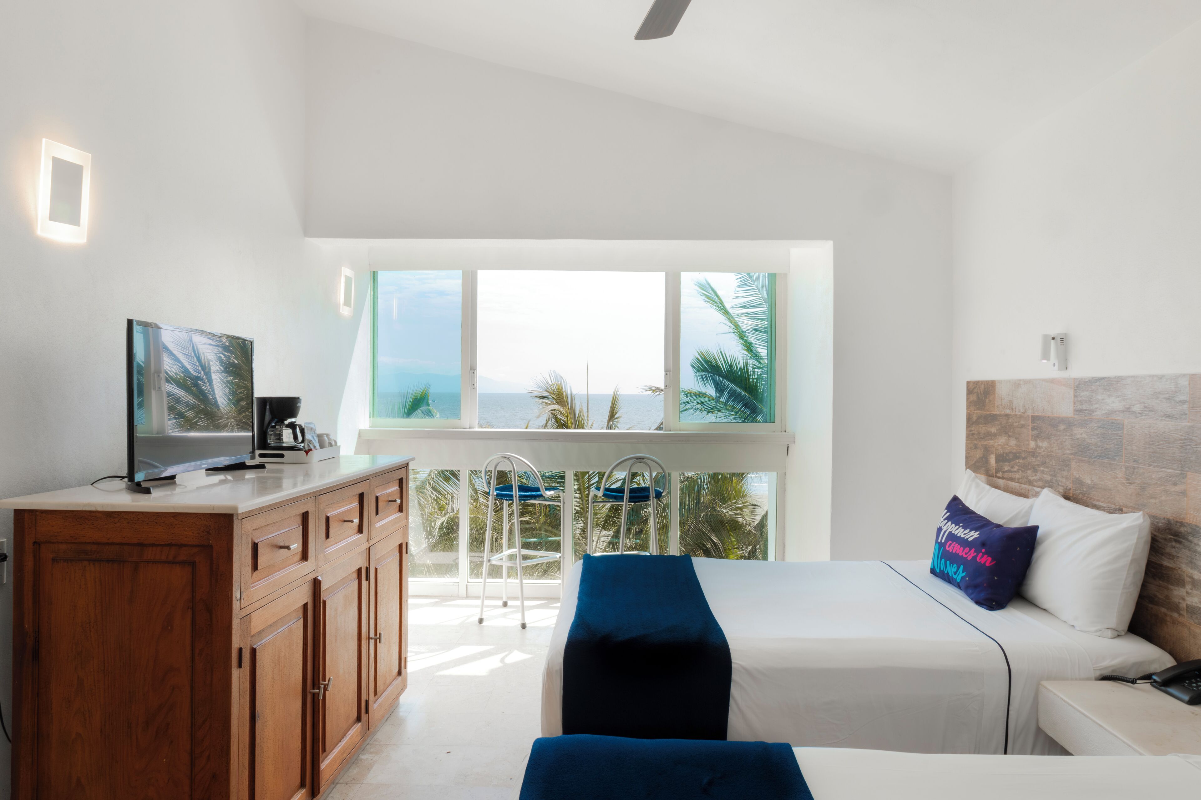 A hotel room with a bed, a TV, a wooden cabinet, and a balcony with a view of the ocean.