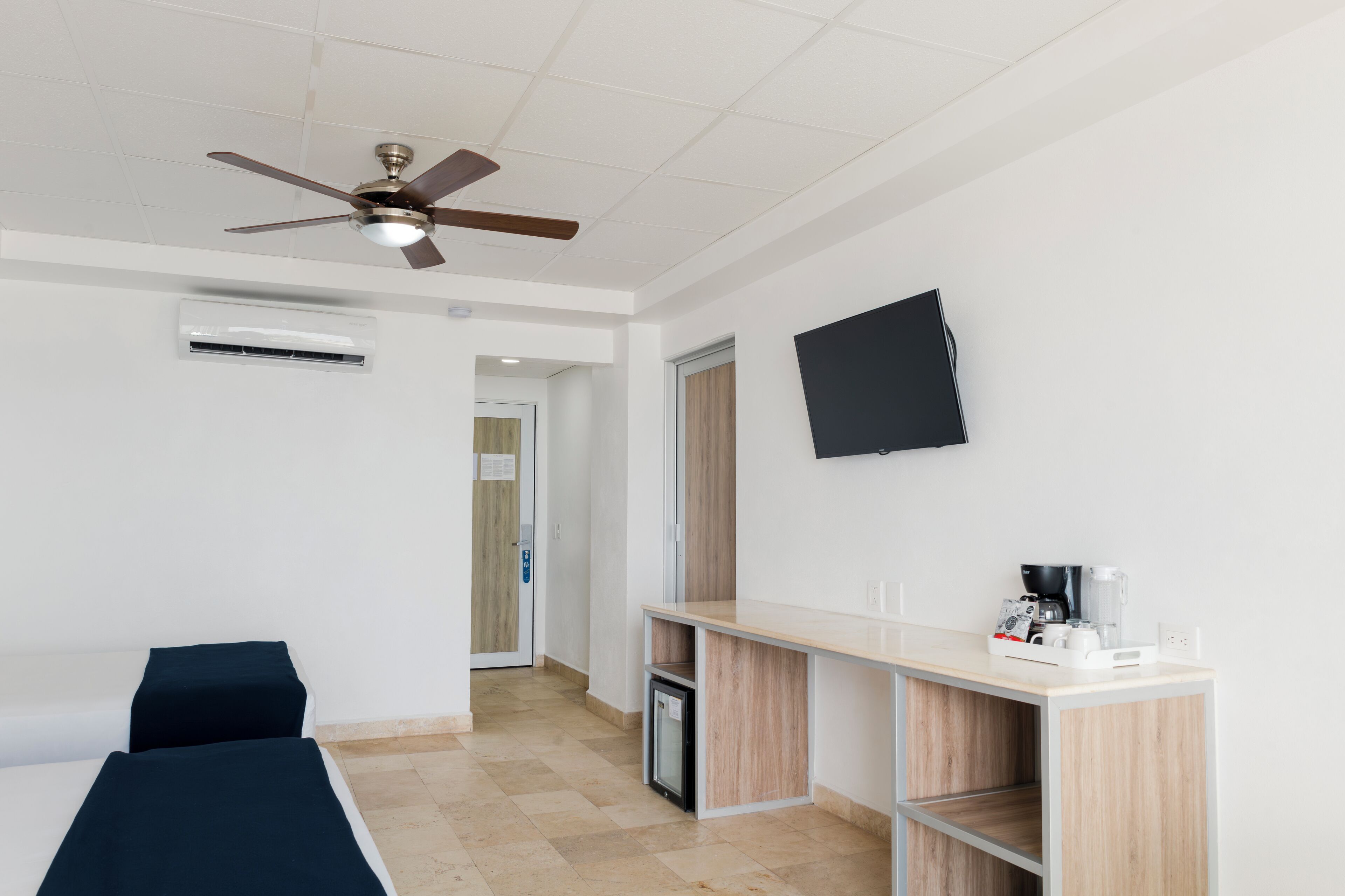 A hotel room with a ceiling fan, a flat-screen TV, a small kitchenette, and a bed with a dark cover.