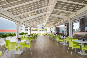 Un área de comedor al aire libre techada con sillas y mesas verdes, piso de madera y vista a edificios al fondo.