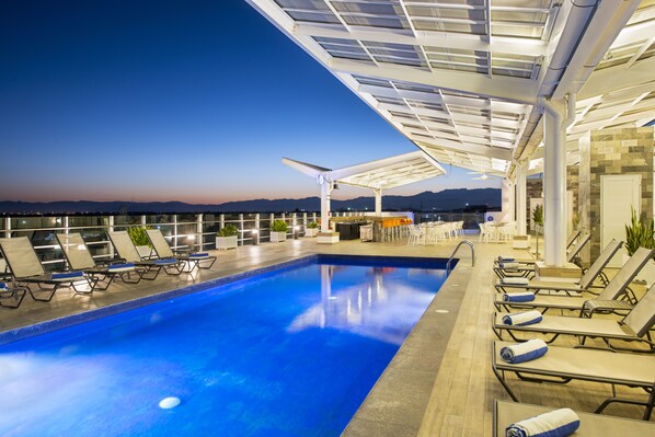 Una piscina en la azotea con sillones de descanso y vistas al horizonte de la ciudad al anochecer.