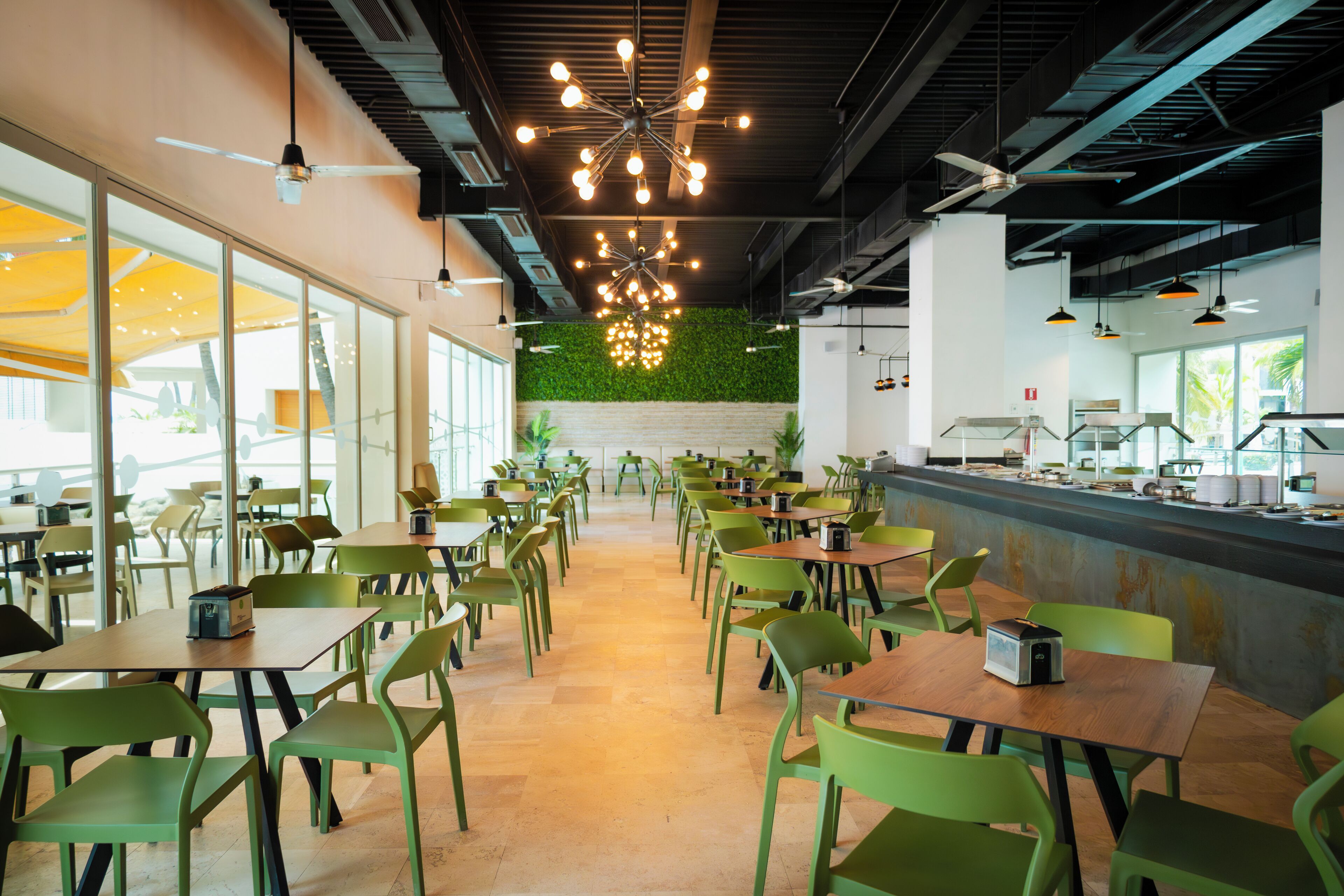 Una cafetería amplia con sillas y mesas verdes, una barra larga con comida y bebidas, y una pared verde con plantas.