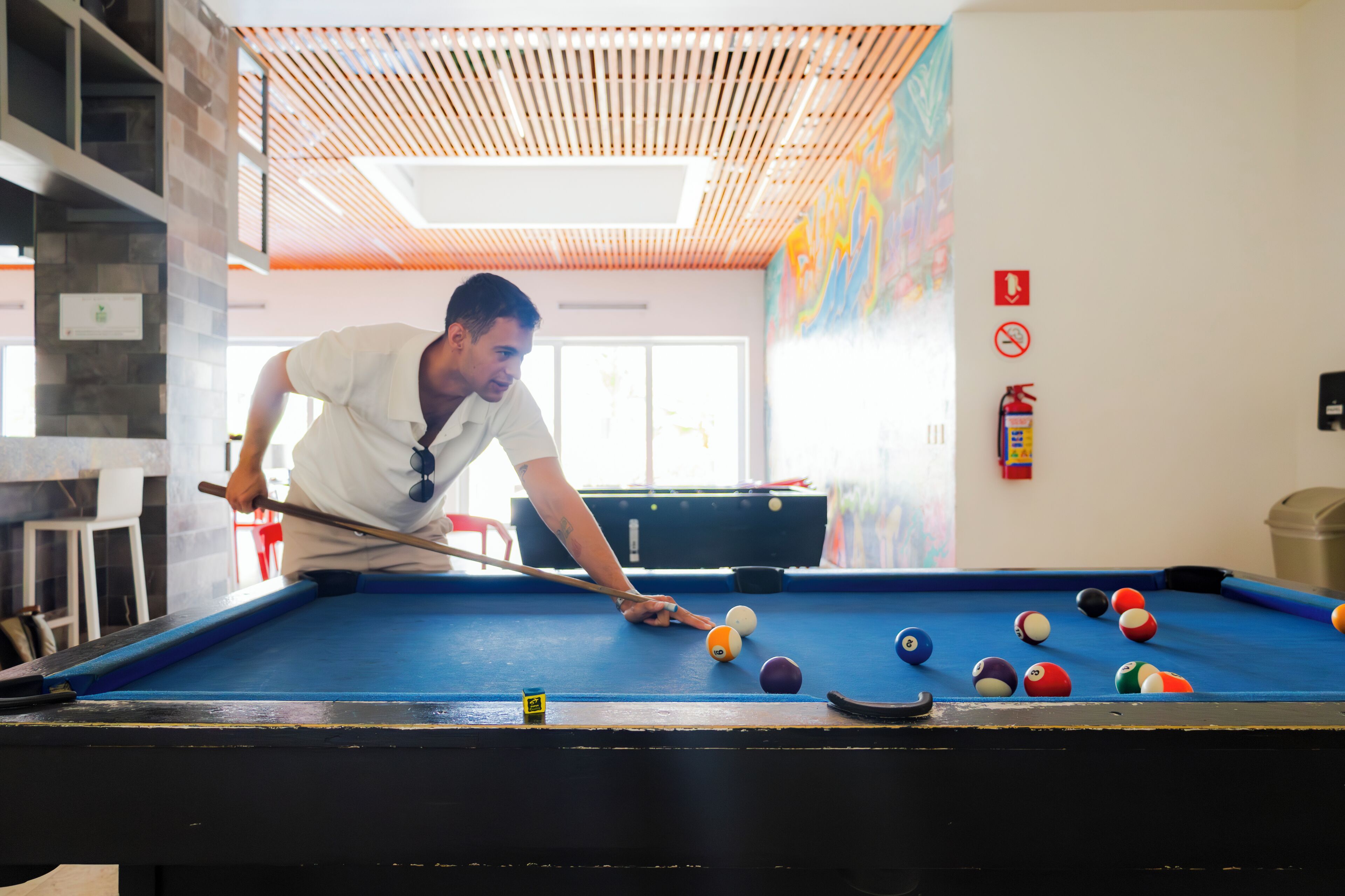 A pool table with a person playing, a fire extinguisher, and a colorful mural on the wall.