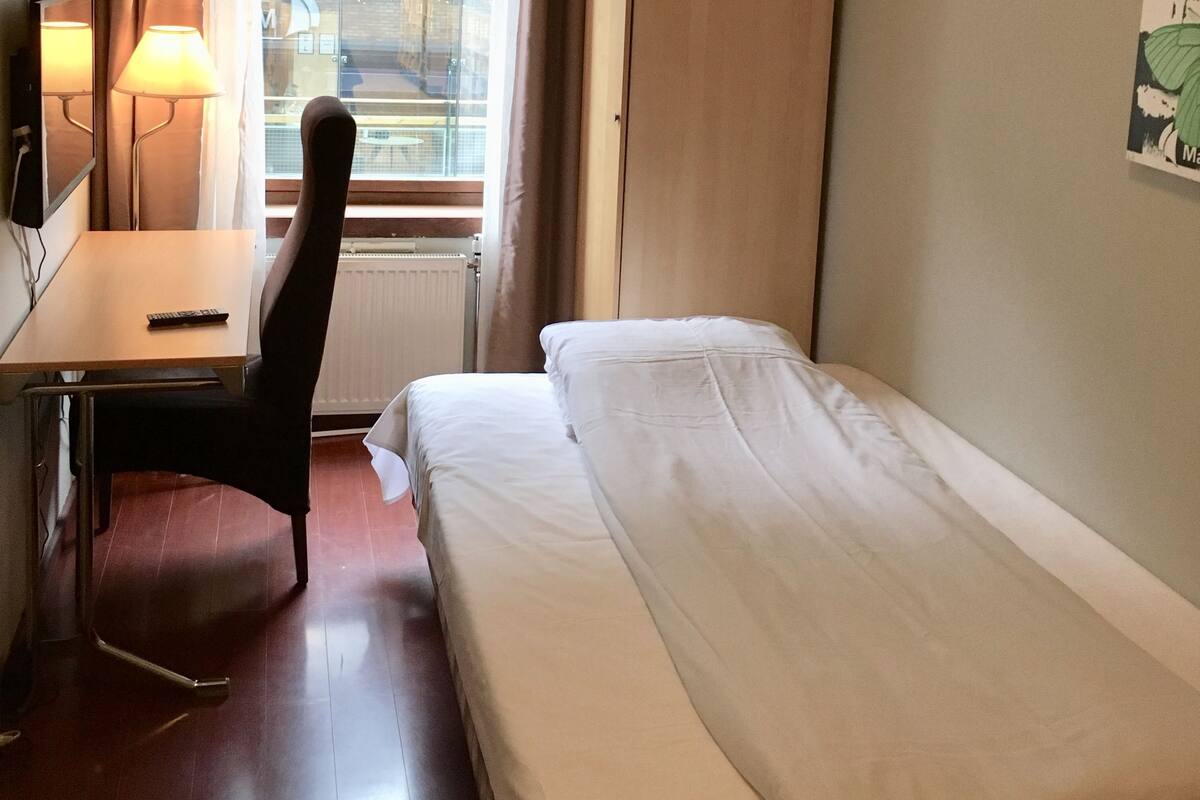 Et hotelværelse med seng, skrivebord, stol, lampe, radiator og et vindue med gardiner.