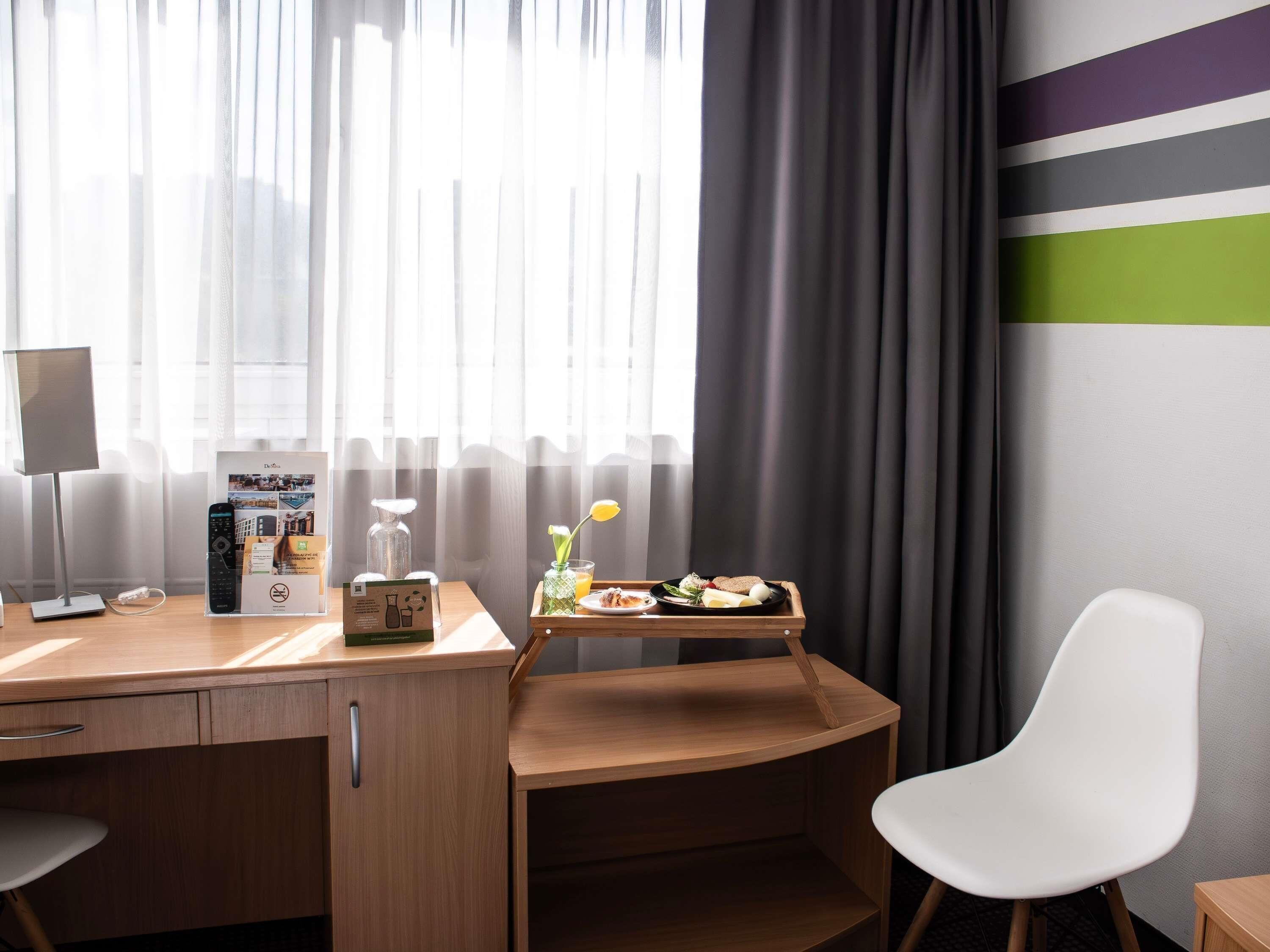 Une chambre d’hôtel équipée d’un bureau, d’une chaise, d’un plateau avec le petit-déjeuner, d’une lampe et d’une fenêtre avec des rideaux.