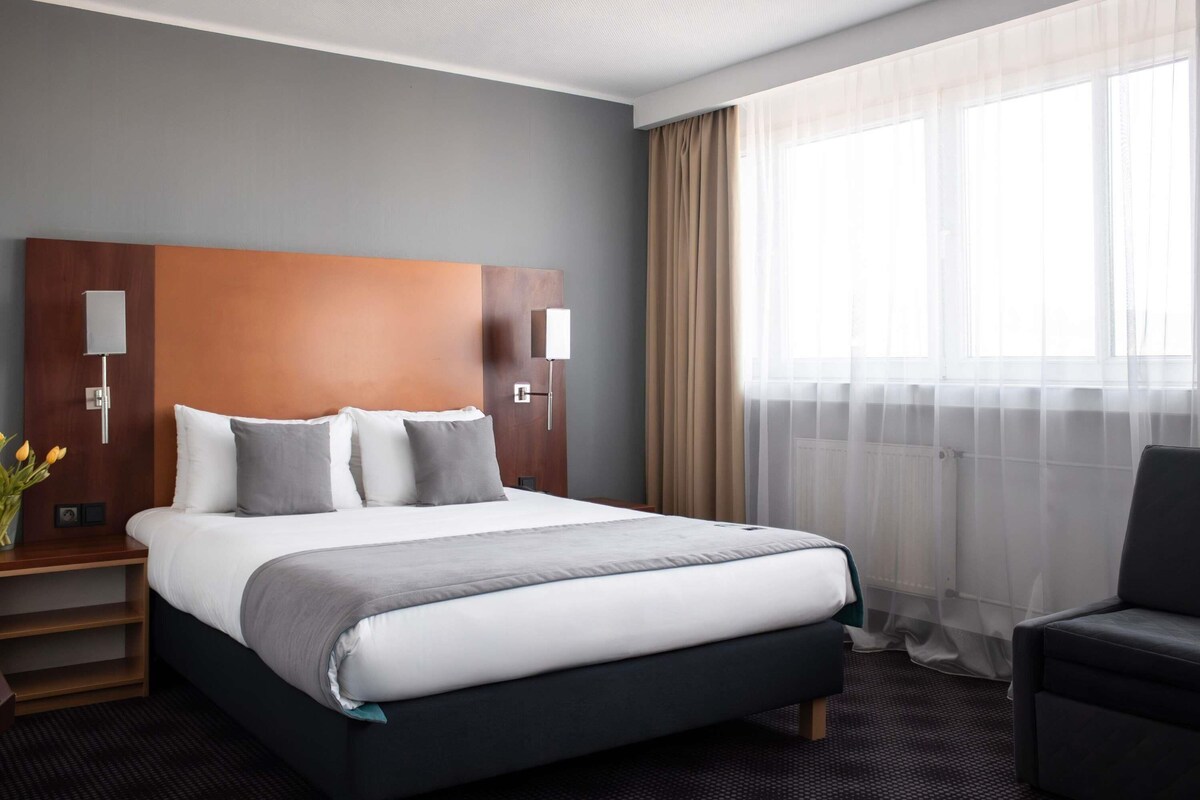 Una habitación de hotel con una cama grande, dos mesitas de noche con lámparas, una silla y una ventana con cortinas de gasa.