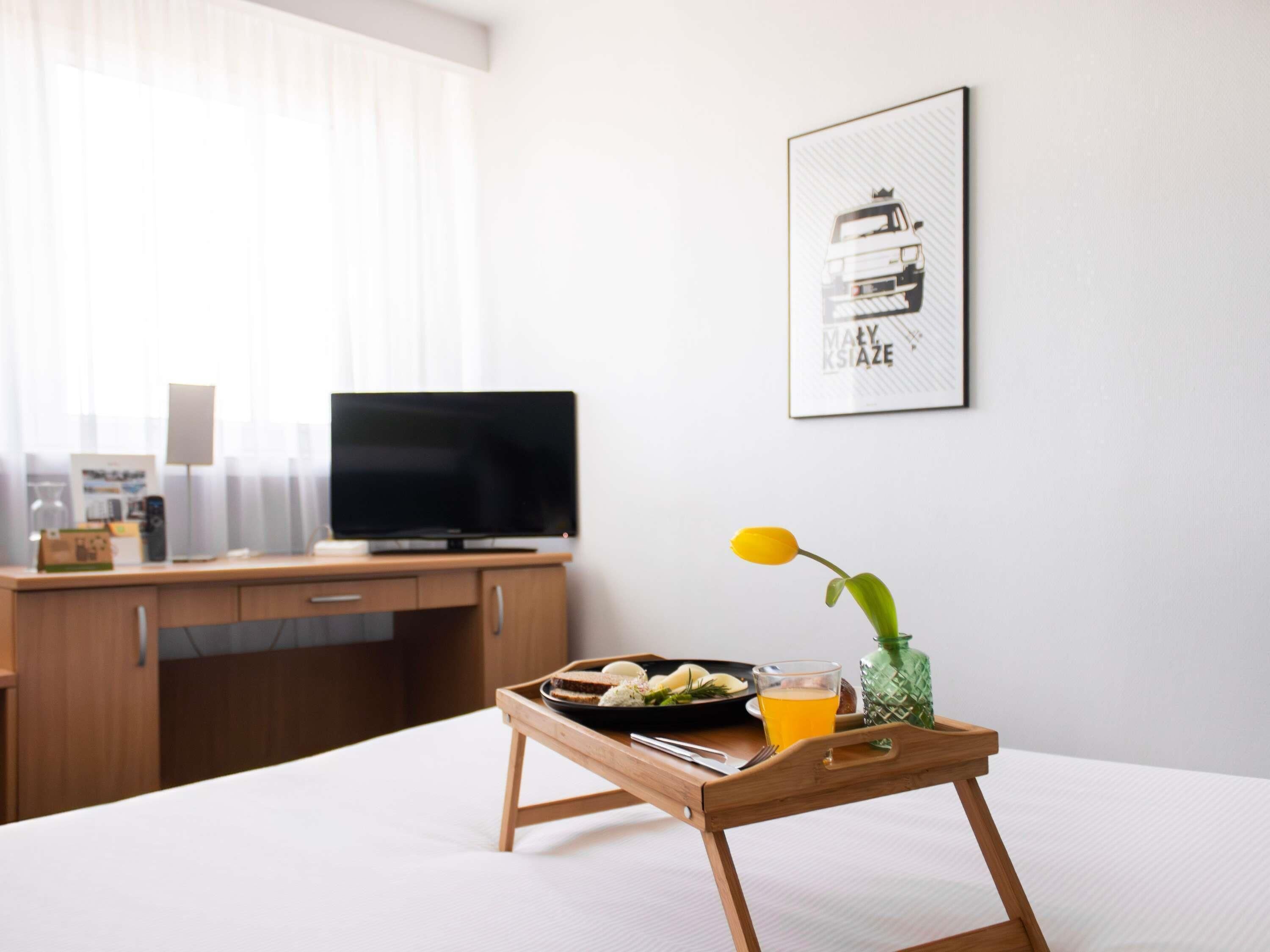 A neatly arranged hotel room with a bed, a bedside table with a tray of breakfast, a television, and a framed picture on the wall.