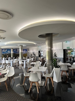 Ein modernes Restaurant mit weißen Tischen und Stühlen, einem polierten Boden und einem Wandbild einer Berglandschaft.