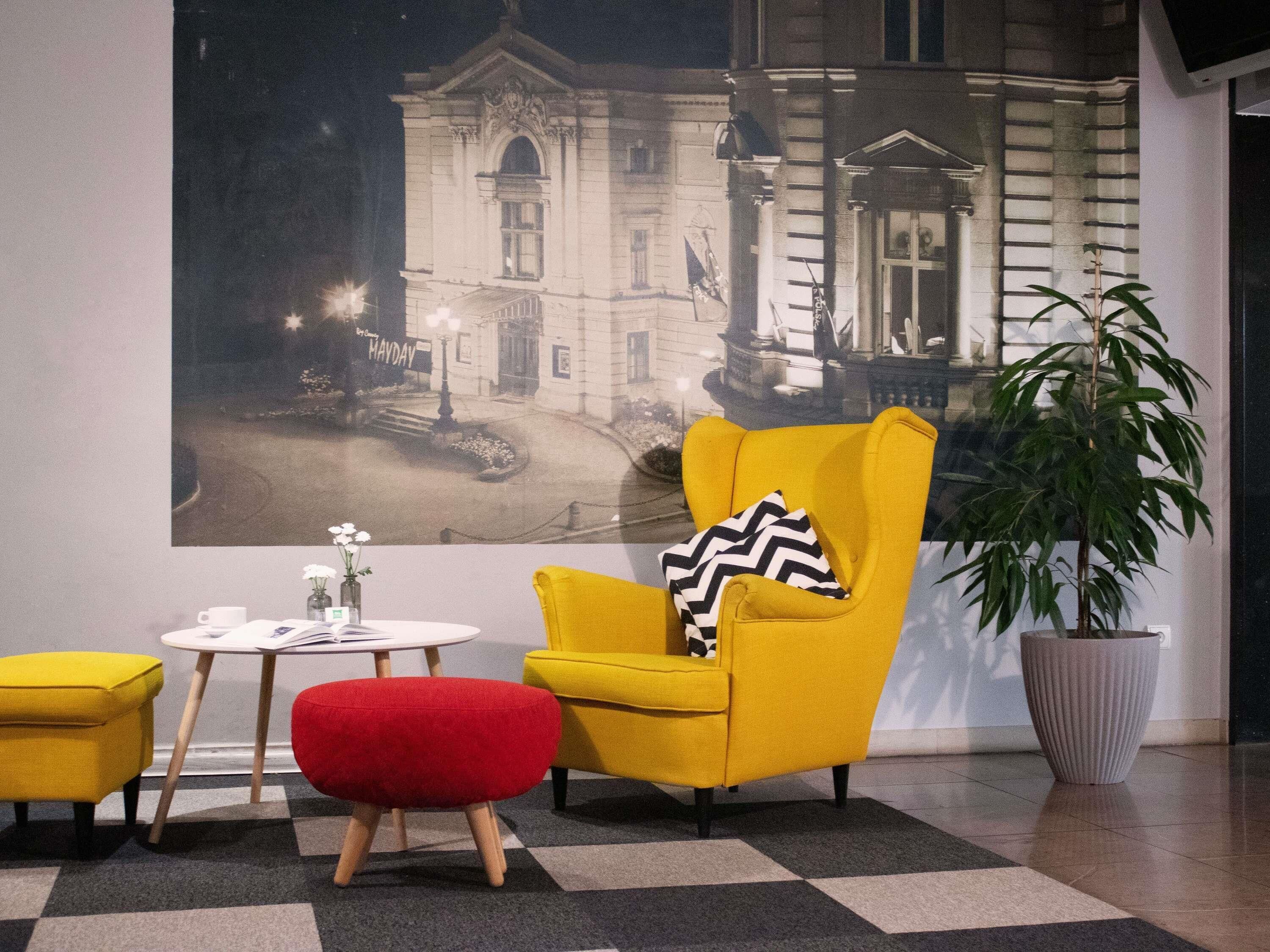 A room with a large poster of a building, a yellow armchair, a red ottoman, a small round table, and a potted plant.