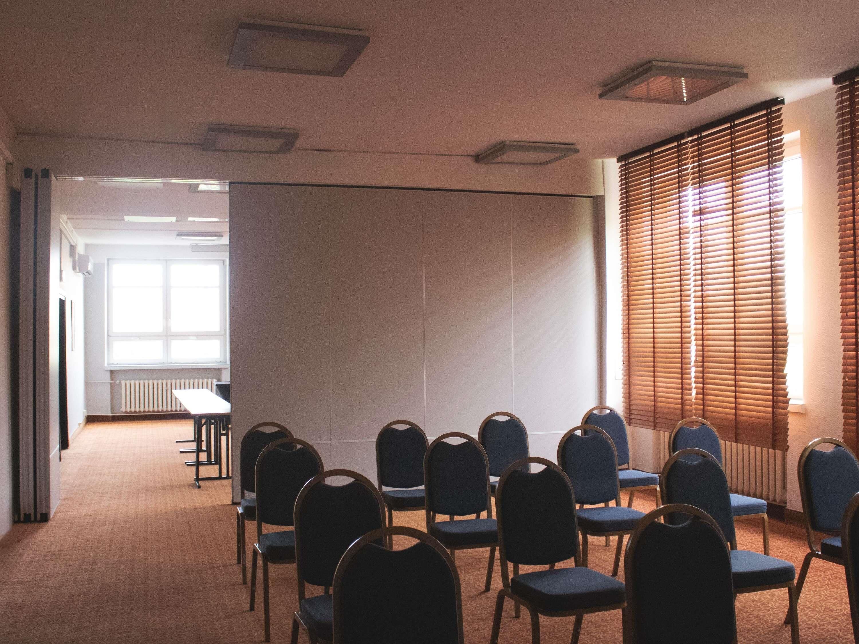 Ein Konferenzraum mit Reihen von Stühlen, einer großen Projektionsleinwand und einem Fenster mit Jalousien.