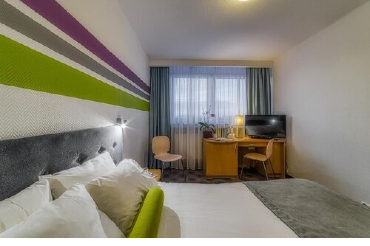 Una habitación de hotel con cama, escritorio, silla, televisor y ventana con cortinas.