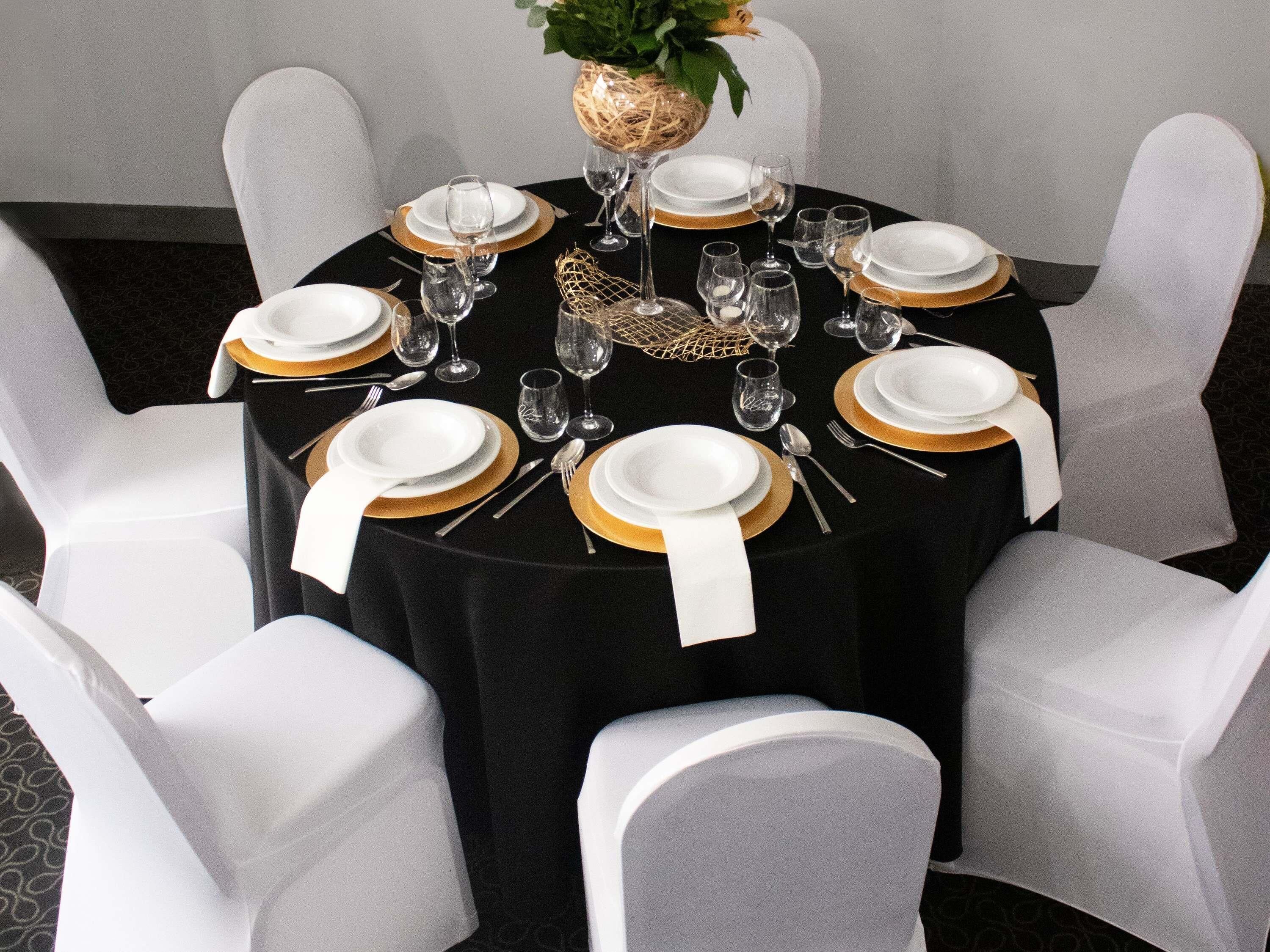A round table set for six with white chairs, black tablecloth, gold accents, and clear glassware.