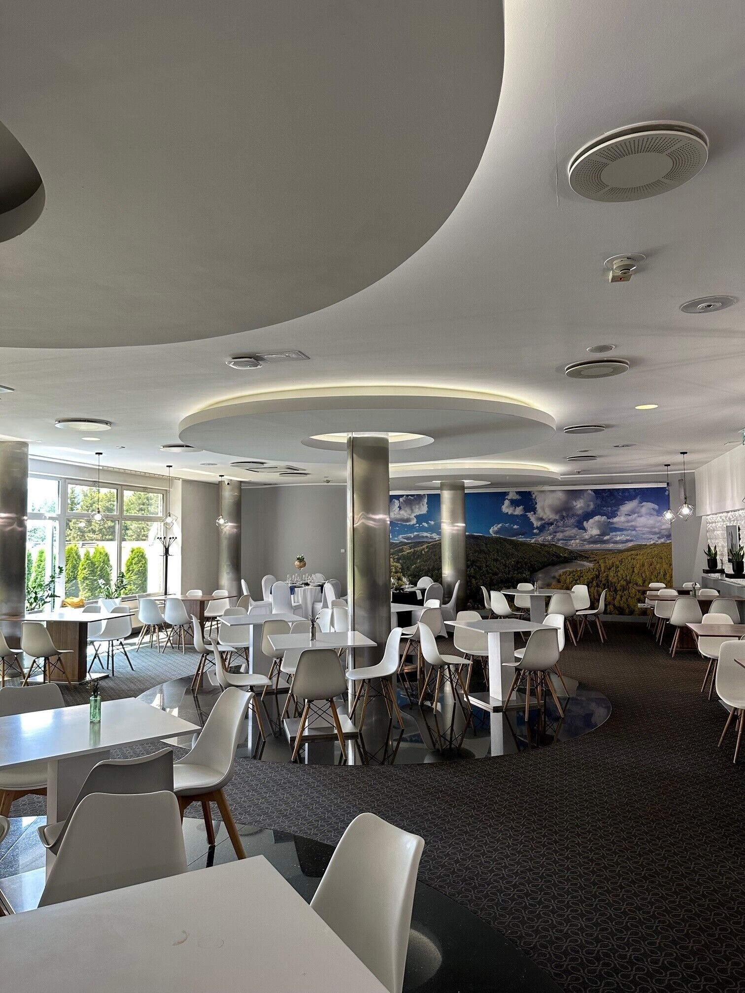 Un comedor moderno con mesas y sillas blancas, ventanas grandes y un mural de un paisaje.