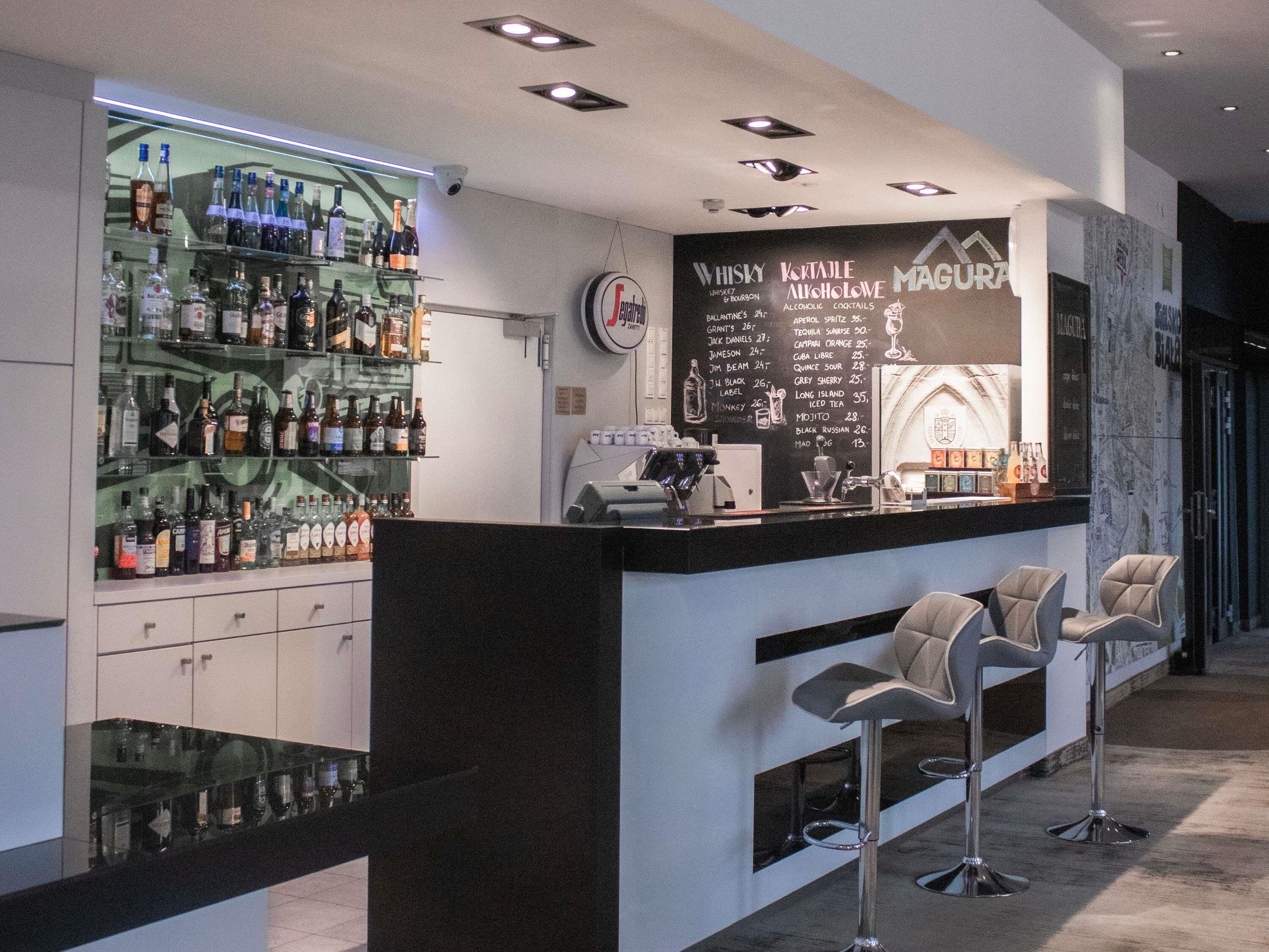 Zona bar con lavagna che mostra un menù di bevande, una vetrina con bottiglie e due sgabelli da bar.