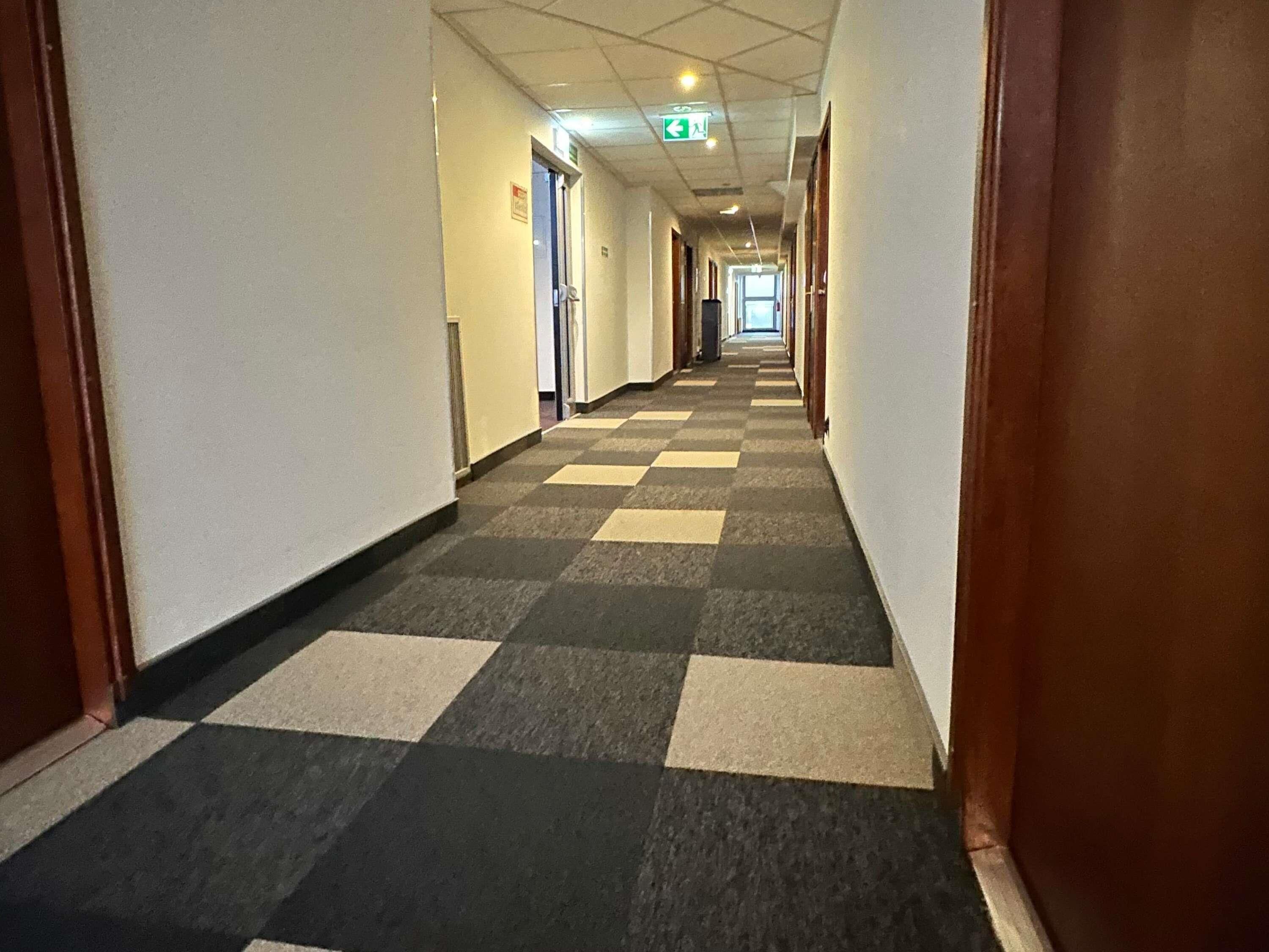 En hotellkorridor med ett rutmönstrat golv, dörrar på båda sidor och en utrymningsskylt i taket.