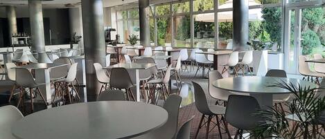 A spacious indoor area with multiple tables and chairs, large windows offering a view of greenery outside, and a modern ceiling design.