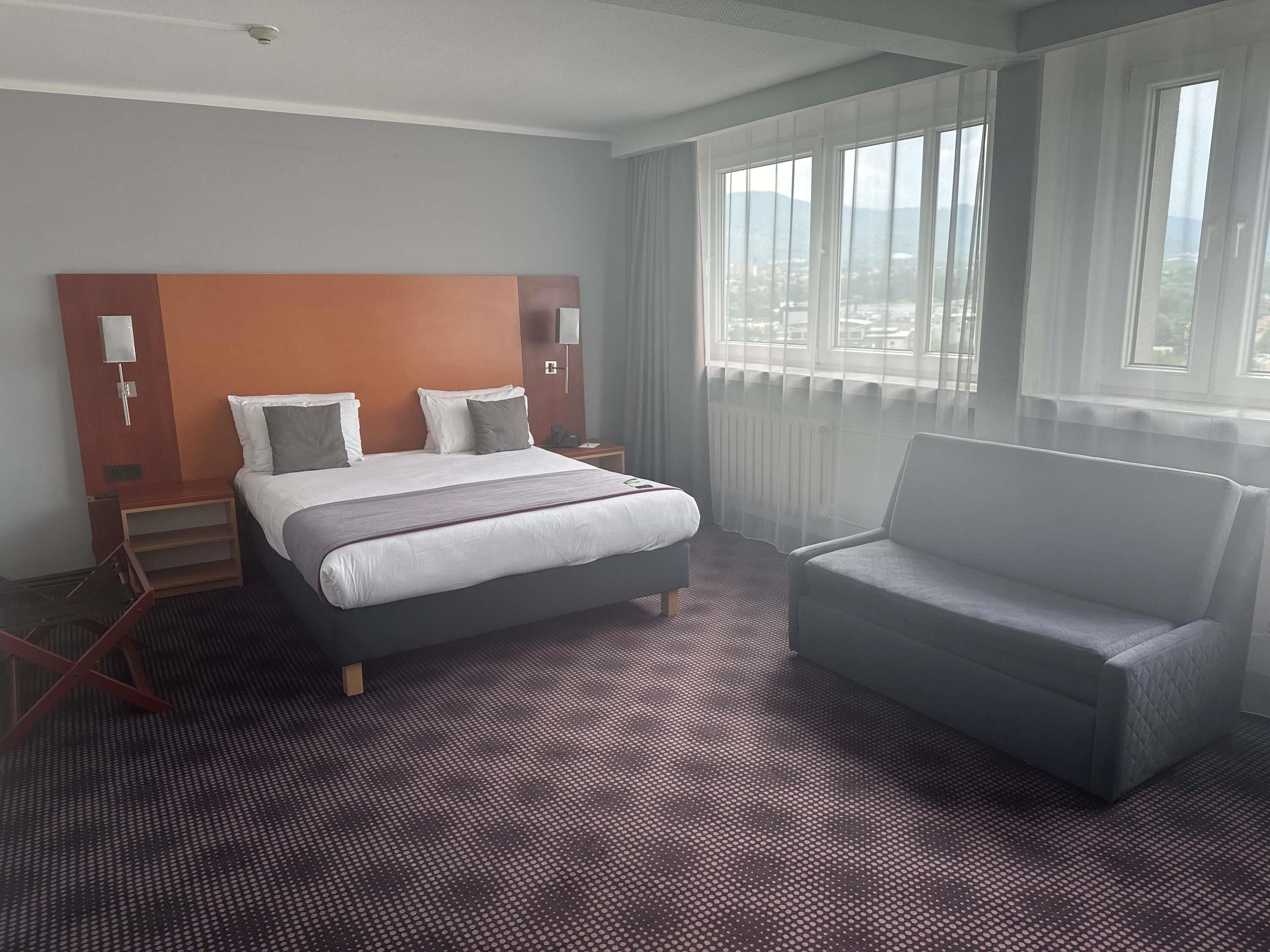 A hotel room with a bed, a chair, a nightstand, and a view of the city through large windows.