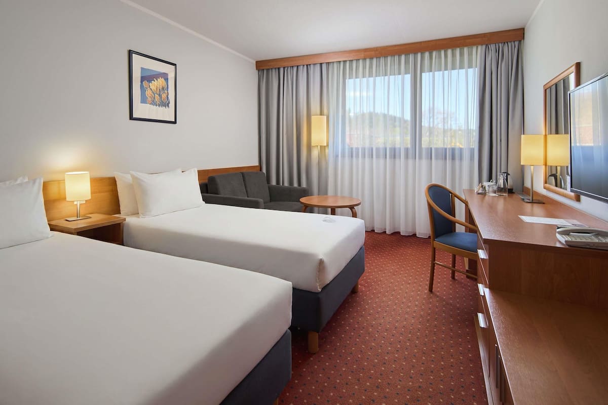 A hotel room with two beds, a desk, a chair, a small table, a lamp, a TV, and a window with curtains.