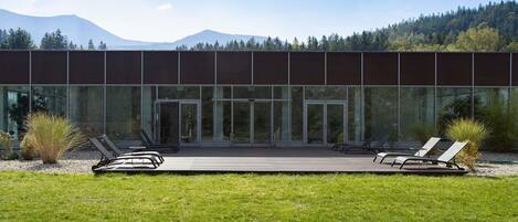 Un edificio moderno con amplios ventanales, una terraza con sillones de descanso y montañas al fondo.