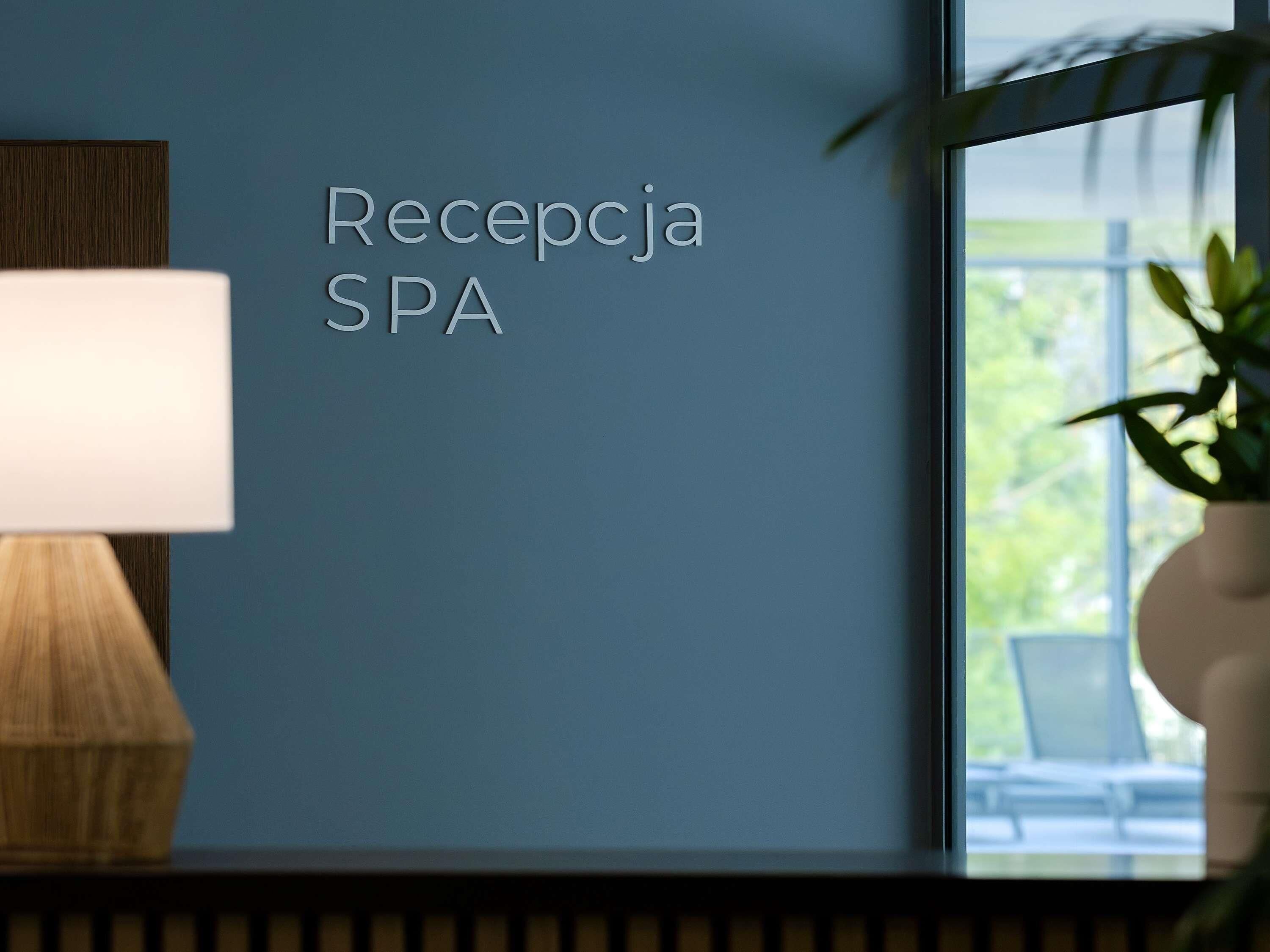 Un área de recepción con un letrero en la pared que dice 'Recepcja SPA' y una lámpara sobre una mesa de madera.