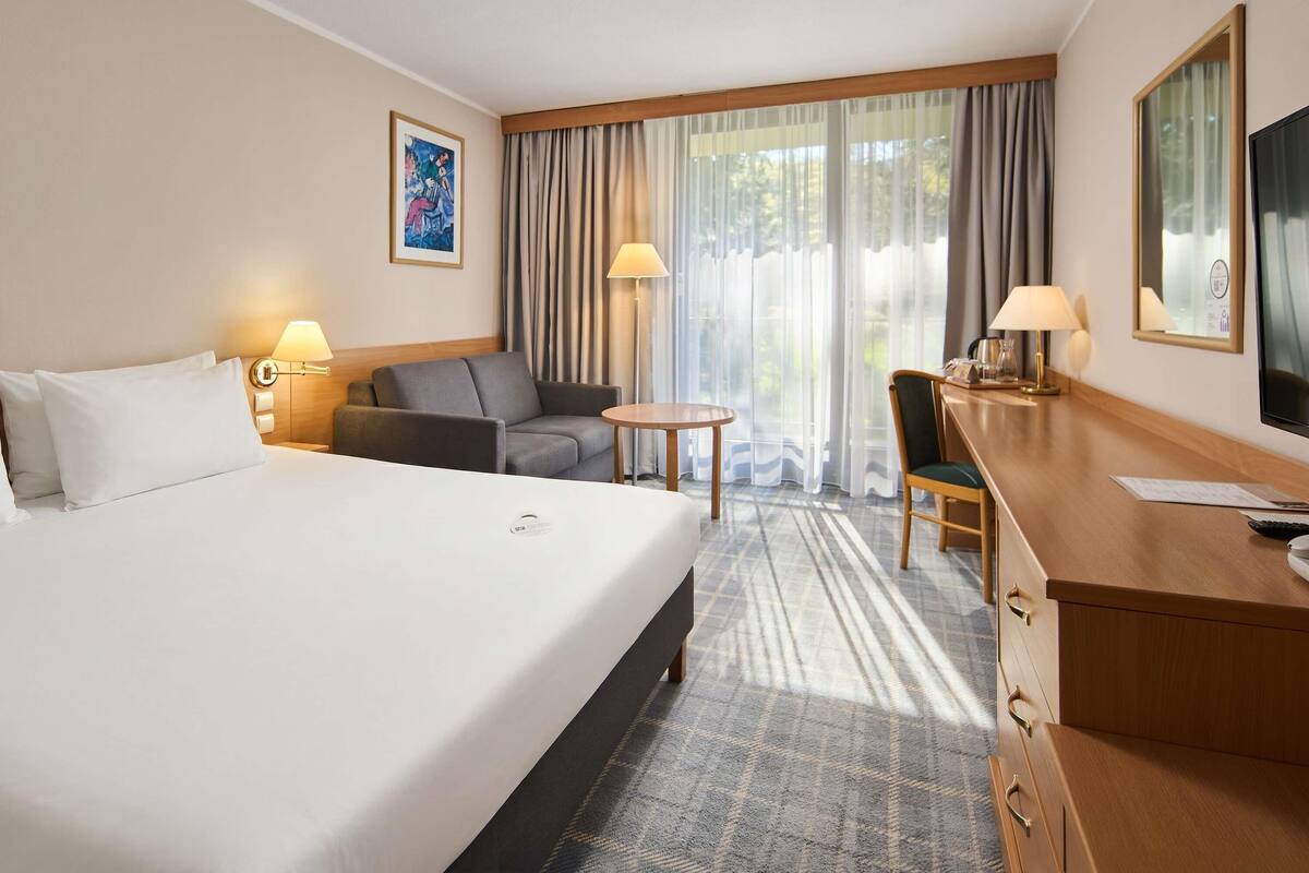 A hotel room with a bed, a sofa, a desk, a chair, a TV, and a window with curtains.