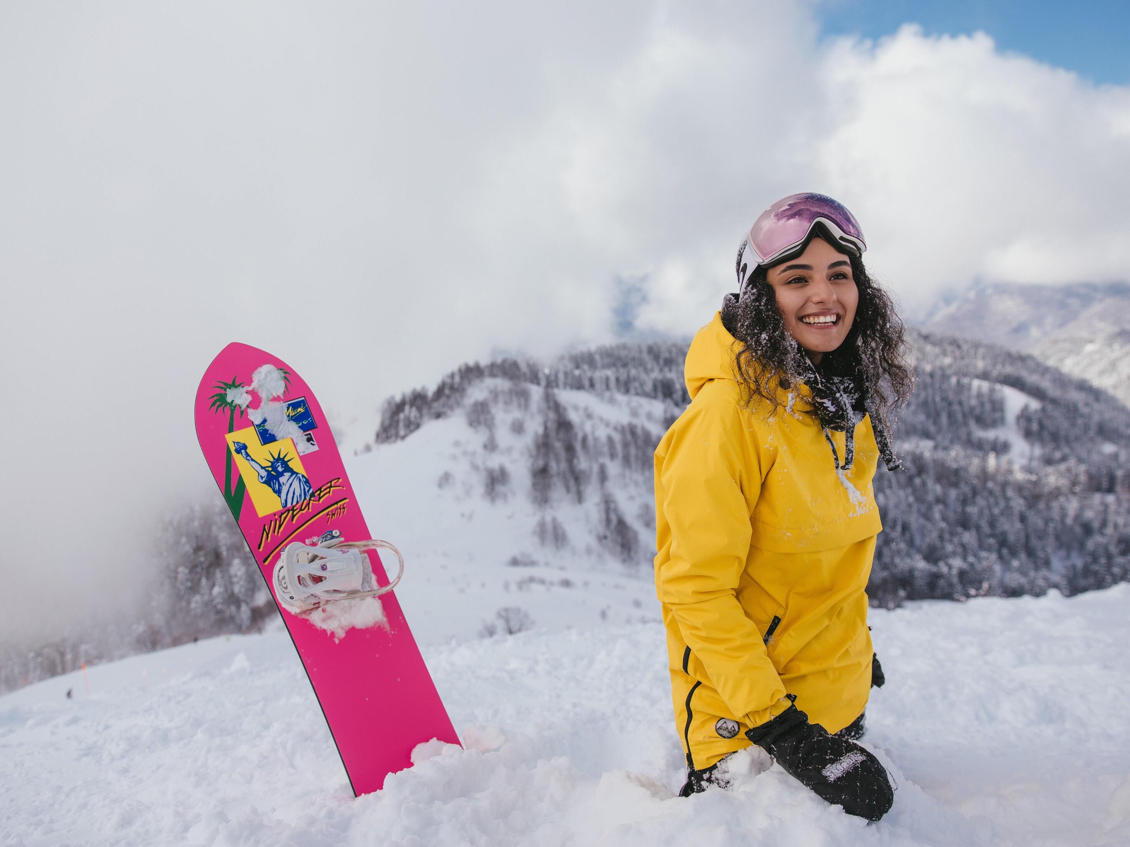 Eine Person mit gelber Jacke und Helm steht neben einem pinken Snowboard. Im Hintergrund ist eine Berglandschaft zu sehen.