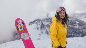 Eine Person mit gelber Jacke und Helm steht neben einem pinken Snowboard. Im Hintergrund ist eine Berglandschaft zu sehen.
