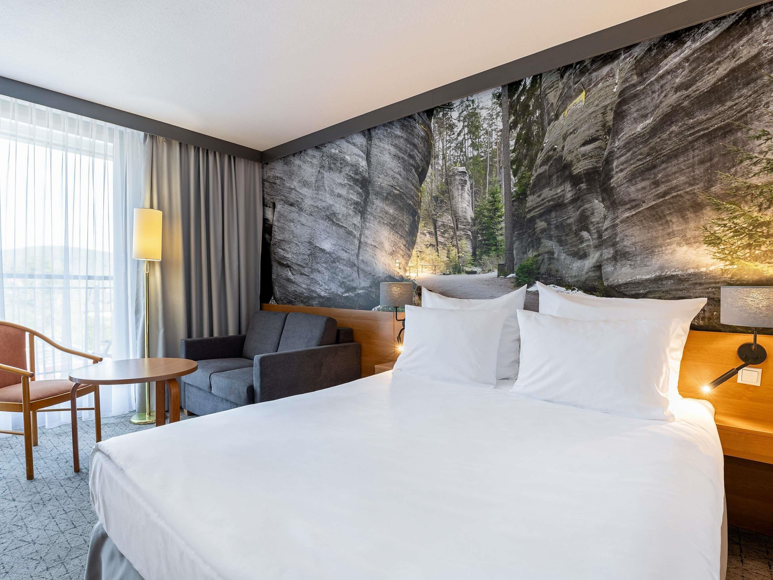 Une chambre d’hôtel avec un grand lit, un coin salon comprenant une chaise et une petite table, une lampe, et une fresque murale représentant une scène de forêt.