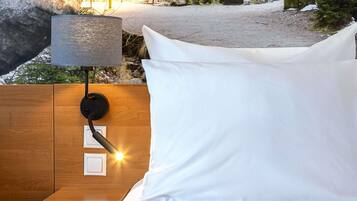 A bed with white bedding, a nightstand, a wall-mounted lamp, and a view of a natural outdoor area.