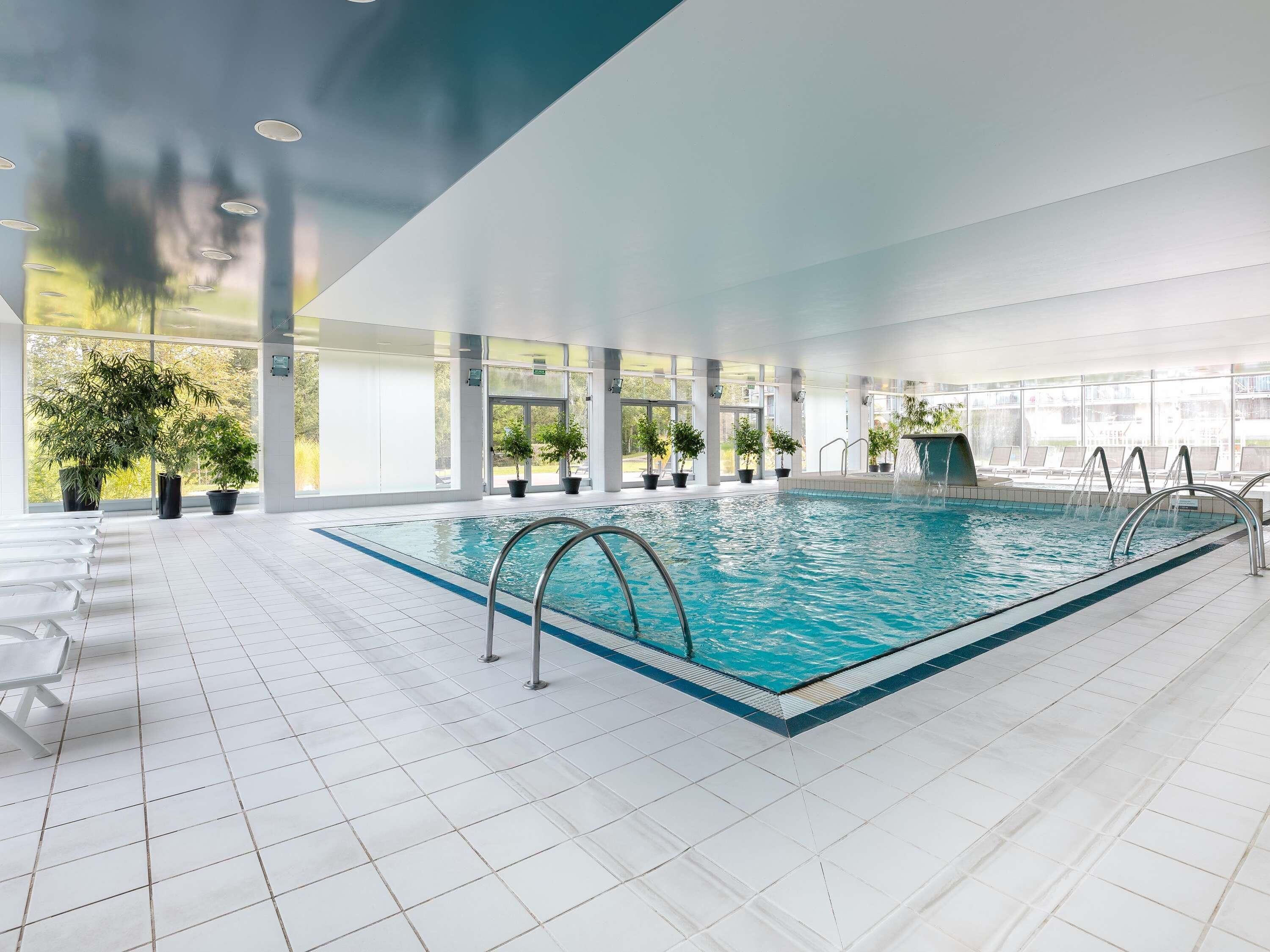 An indoor swimming pool with clear blue water, surrounded by white tiled floors and walls, with potted plants and a waterfall feature.
