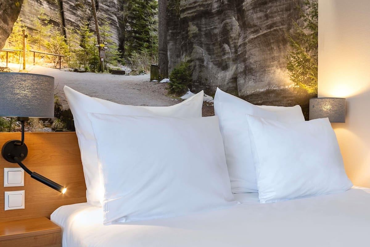 A bed with white bedding, a nightstand with a lamp, and a wall-mounted light fixture.
