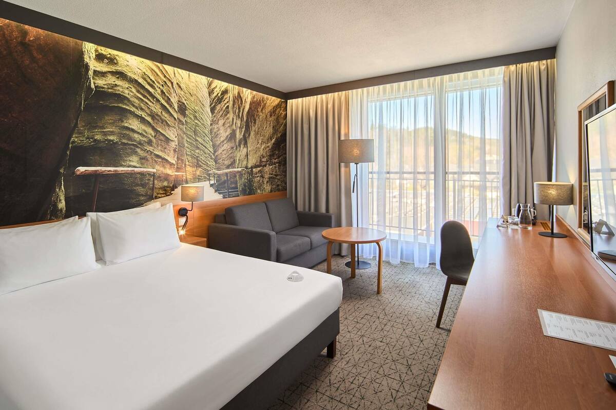 A hotel room with a large bed, a sofa, a small table, a desk, and a large window with curtains.