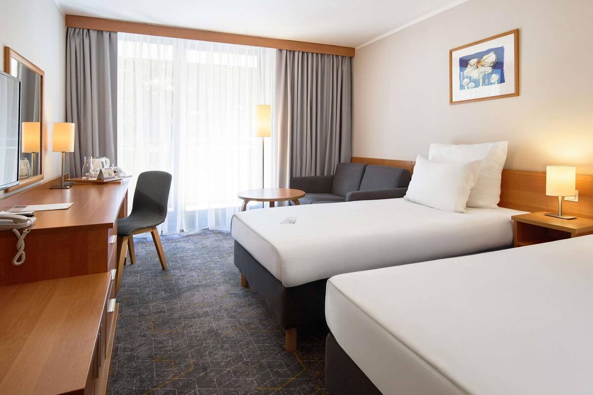 A hotel room with a desk, two beds, a chair, a small table, and a lamp.
