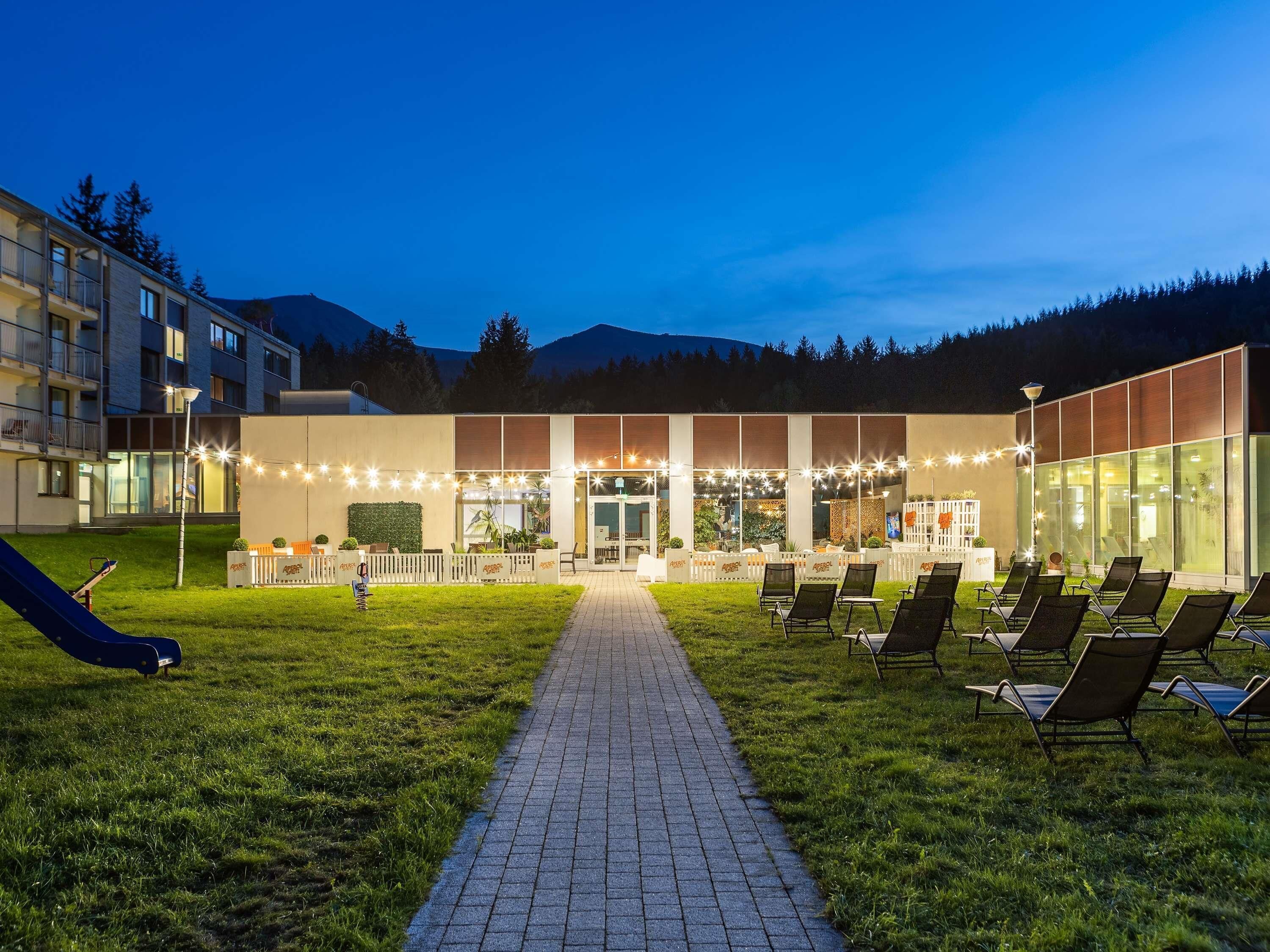 A building with a paved walkway leading to a seating area with chairs and tables, surrounded by greenery and a mountain backdrop.