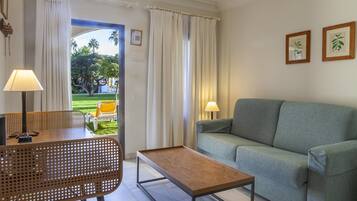 A living room with a sofa, a wooden coffee table, a wicker chair, and two lamps. There is a view of a garden through a doorway.