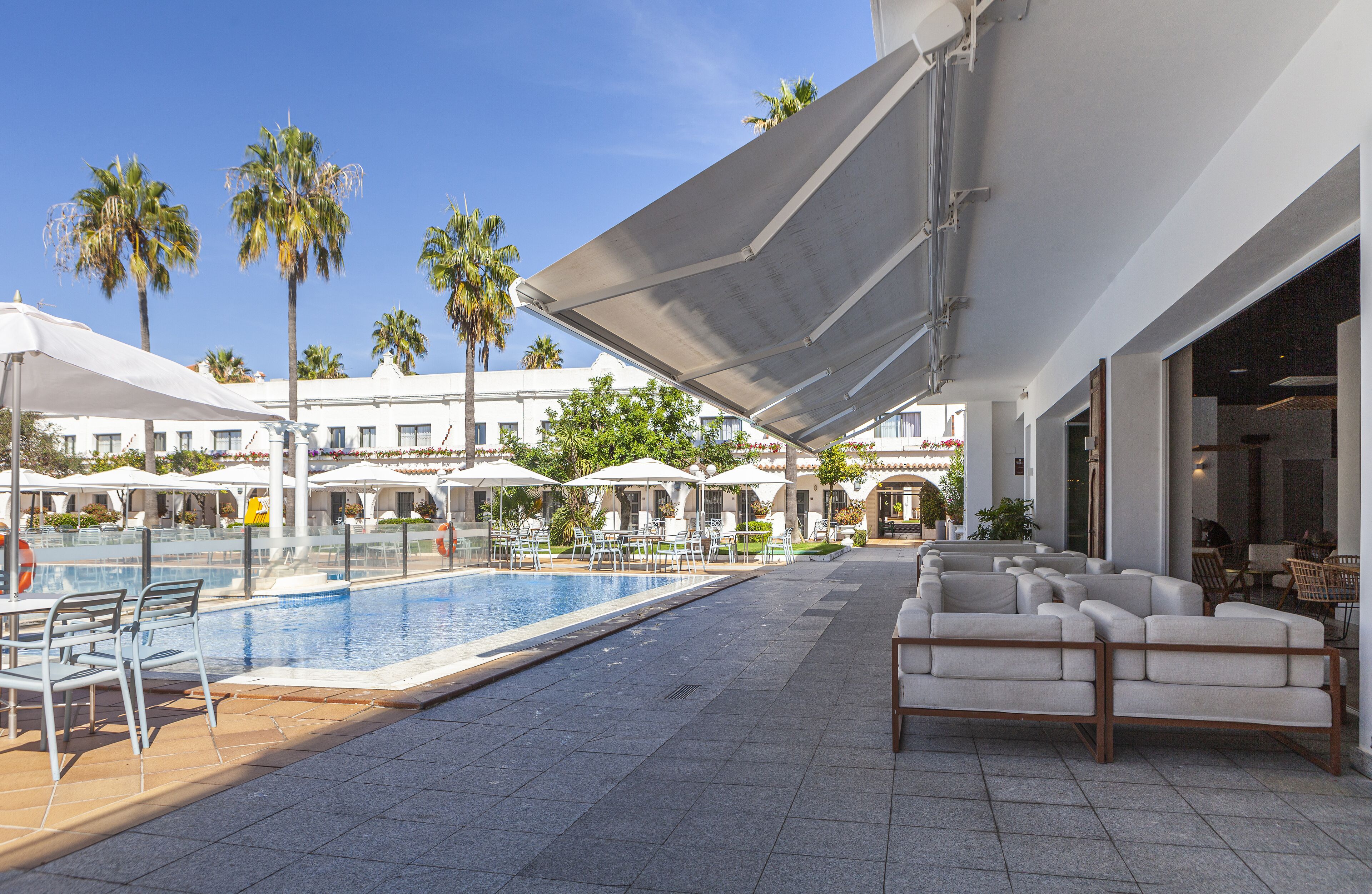 A pool area with lounge chairs and umbrellas, palm trees, and a building with large windows.