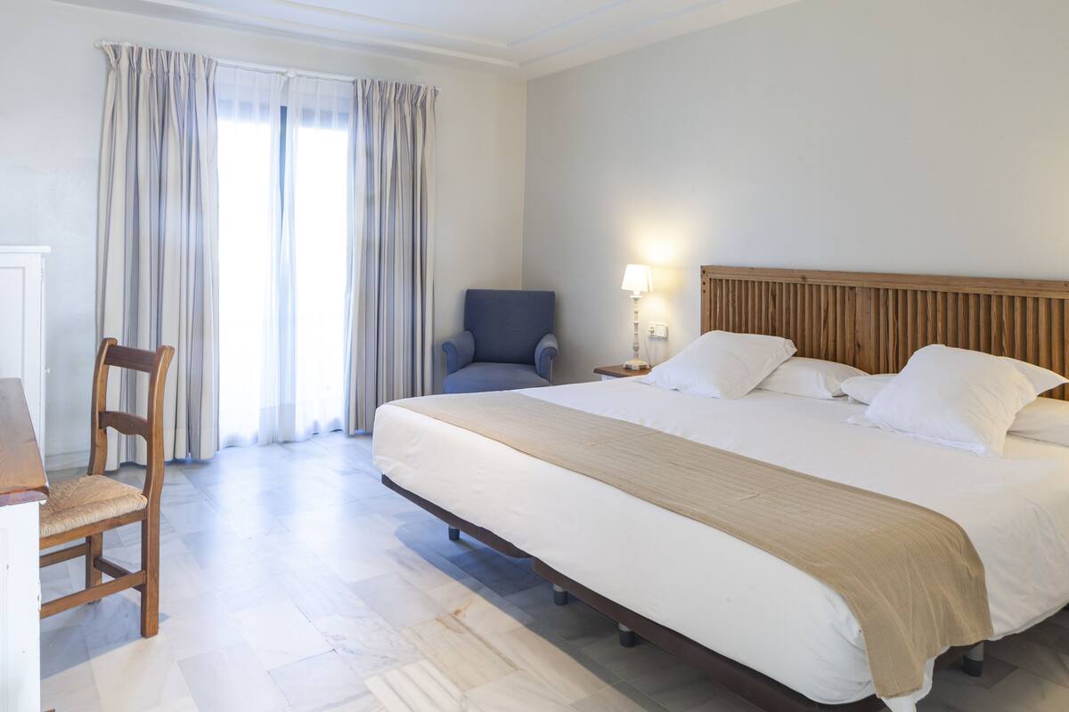Une chambre d’hôtel avec un grand lit, une chaise en bois, un bureau et une lampe.