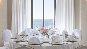 Una mesa de comedor dispuesta para una comida, con manteles blancos, platos, vasos y cubiertos, ubicada frente a un ventanal con vistas al mar.