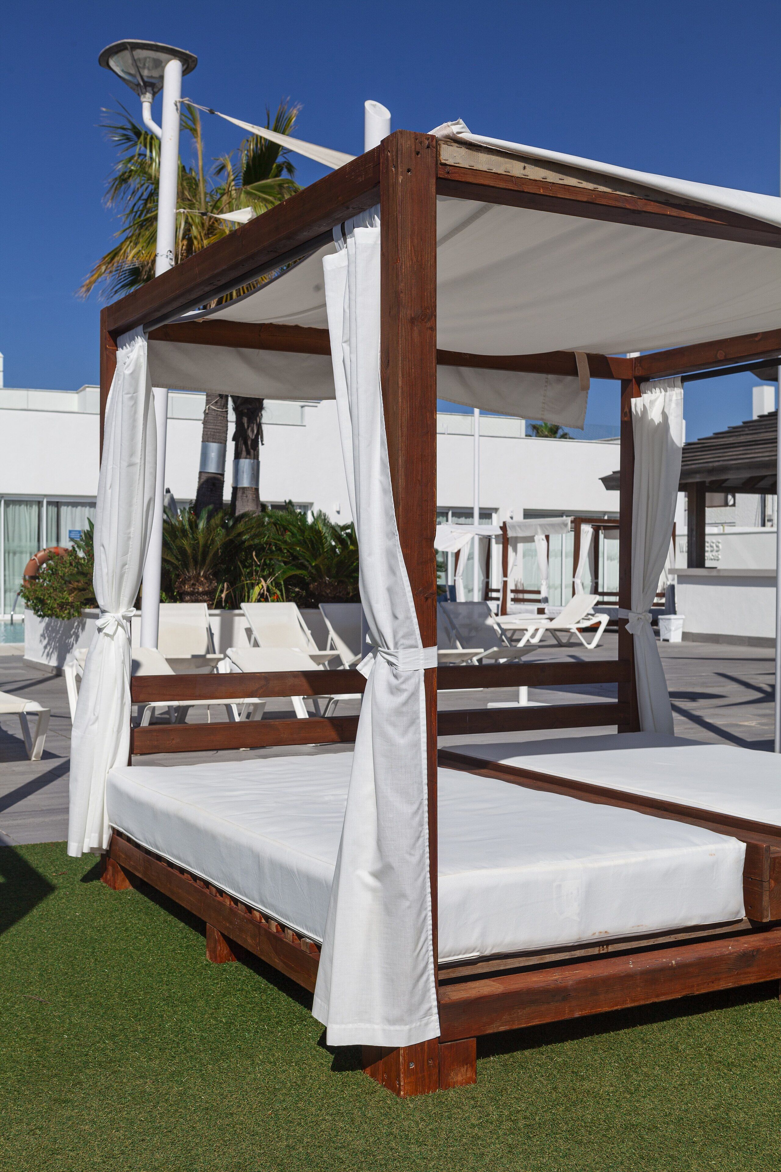 A wooden four-poster bed with white linens, surrounded by white chairs and a small potted plant.