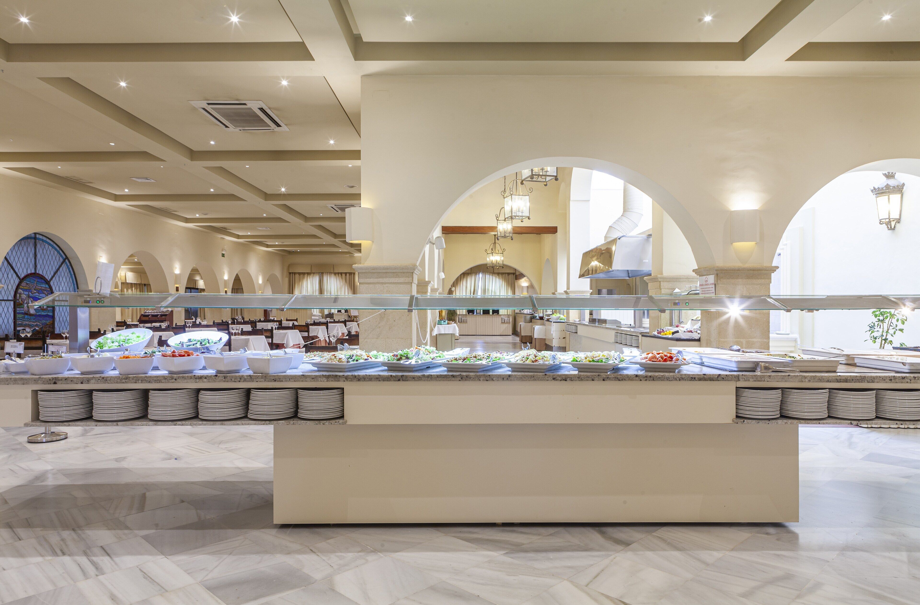 Uma área de buffet com várias estações de comida, piso de mármore e teto alto com iluminação embutida.